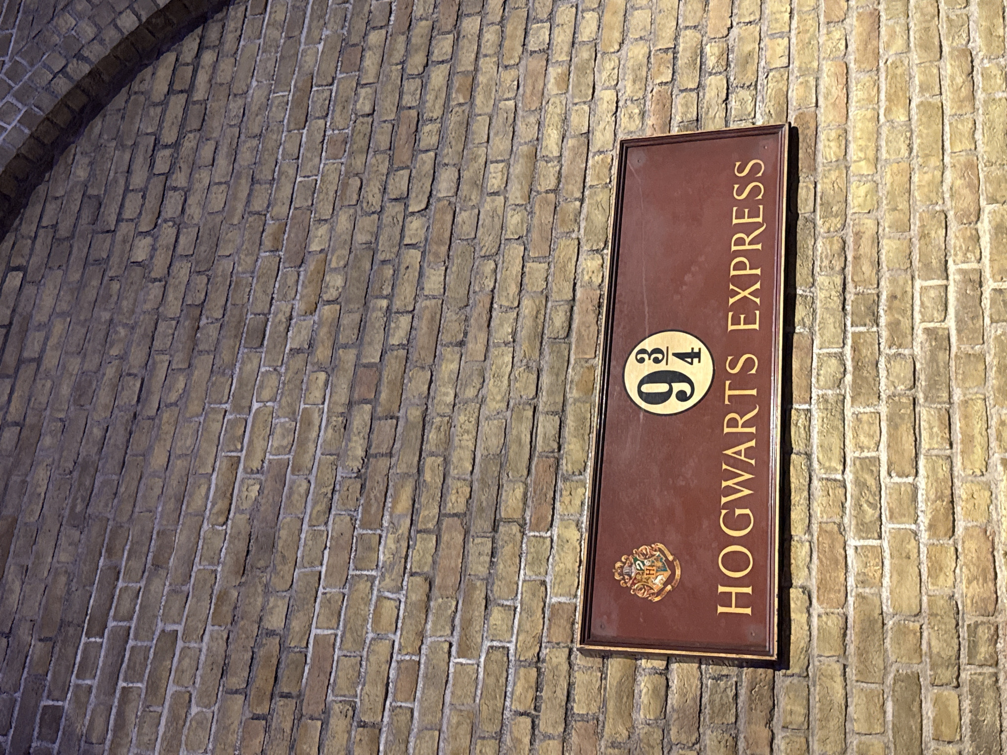A sign that reads "HOGWARTS EXPRESS" is mounted on a brick wall. The sign is rectangular with a dark red background and gold lettering. To the left of the text is an emblem with a shield and a crest. To the right of the emblem is a circular emblem with the number "9 3/4" in gold. The brick wall appears to be made of light brown bricks with white mortar. The sign is positioned at an angle, suggesting it may be part of a larger display or attraction. The background is a brick wall that curves upwards, possibly forming an arch. The lighting suggests an indoor setting. There are no people visible in the image. The location is Platform 9 ¾ at King's Cross Station in London, United Kingdom.