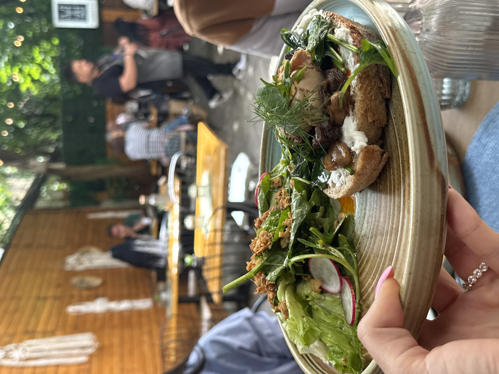 A medium shot captures a person's hand holding a plate of food at an outdoor restaurant in an unknown city. The plate contains a salad with lettuce and radish slices, and an open-faced sandwich topped with cream cheese, mushrooms, and greens. In the background, blurred patrons are seated at tables, and greenery from overhead trees dapples the scene with light. The wall behind the tables is adorned with macrame wall hangings.