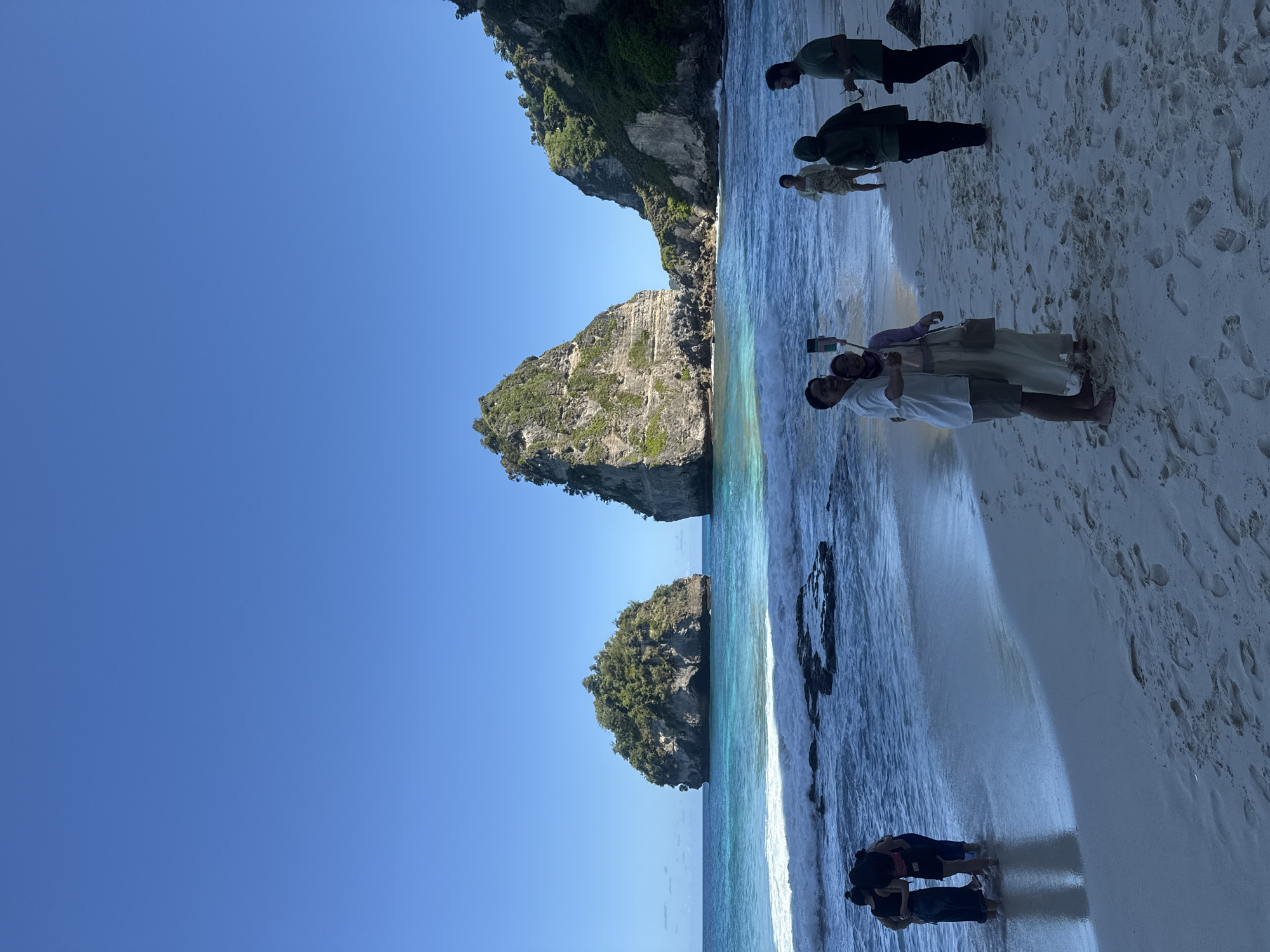 The image depicts a beach scene in Nusa Penida, Indonesia, with a clear blue sky. In the distance are two large, rocky, tree-covered islands. The ocean water is turquoise. The sandy beach is bright. Several groups of people are present. One group of three appears to be taking a selfie. A second group is standing near the water's edge. A third group is further up the beach.
