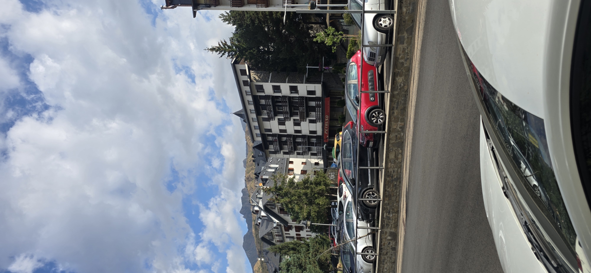 The image shows a street scene in Sallent de Gállego, Spain, with a row of parked cars along a stone wall. Behind the cars, several multi-story buildings with balconies are visible, some of which appear to be hotels or apartment complexes. Trees are interspersed among the buildings, and in the background, mountains rise against a partly cloudy sky. The street in the foreground is paved, and the hood of a white car is visible at the bottom of the frame, suggesting the photo was taken from within another vehicle. The overall impression is of a mountain town on a pleasant day.