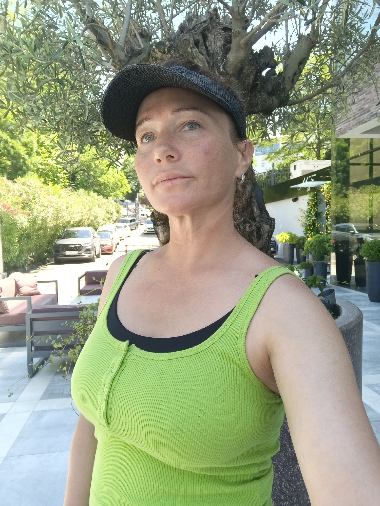 A medium shot photograph captures a woman standing outdoors, positioned slightly to the right of the frame. She is facing the camera, looking upwards and to her left. The woman is wearing a bright green ribbed tank top layered over a black bra, and a dark grey baseball cap. Her facial expression is neutral, and she has fair skin with freckles. Behind her, a large tree with intricate branches dominates the upper portion of the image, partially obscuring the background. In the middle ground, a row of parked cars is visible on a street, with lush green foliage lining the left side of the road. A modern building with large glass windows and an illuminated facade can be seen on the right side of the background, with potted plants placed outside. The ground in the foreground is paved with light-colored tiles. The lighting suggests it is daytime, likely sunny. The scene appears to be set in a somewhat urban or suburban environment, possibly a street in Sochi, Russia, given the presence of Mediterranean-style vegetation and architecture.