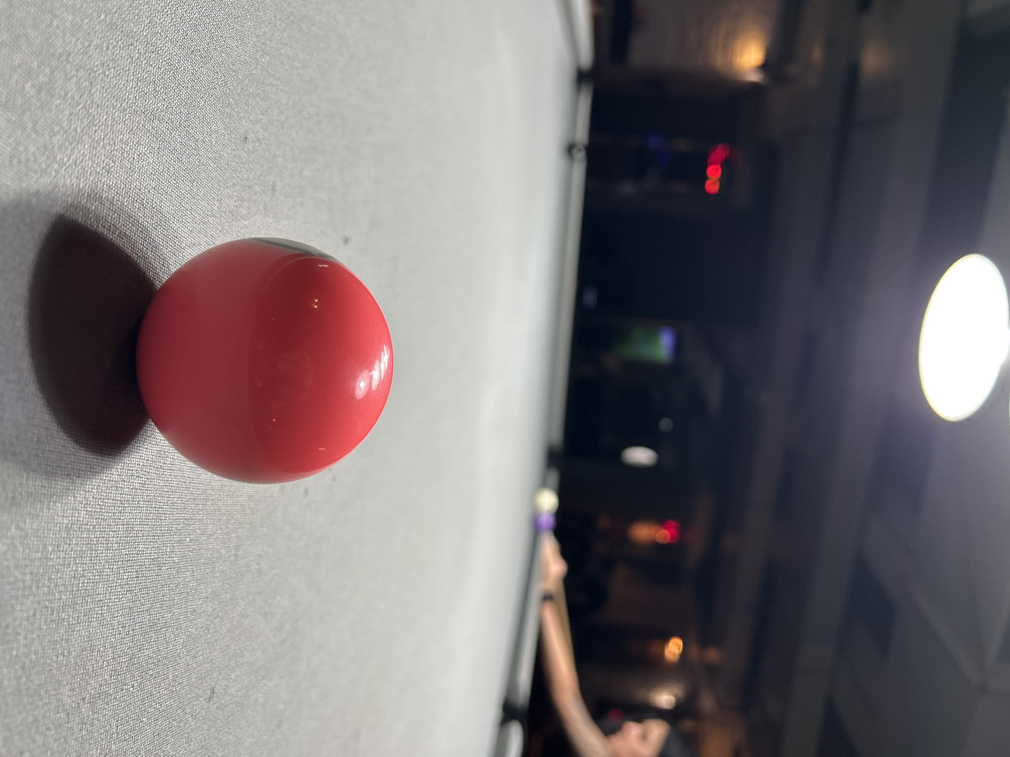A red pool ball sits on a gray pool table. In the blurred background is a dimly lit room with a person playing pool. Red and orange lights are visible in the distance, along with a bright overhead light. The exact location cannot be determined from the image.