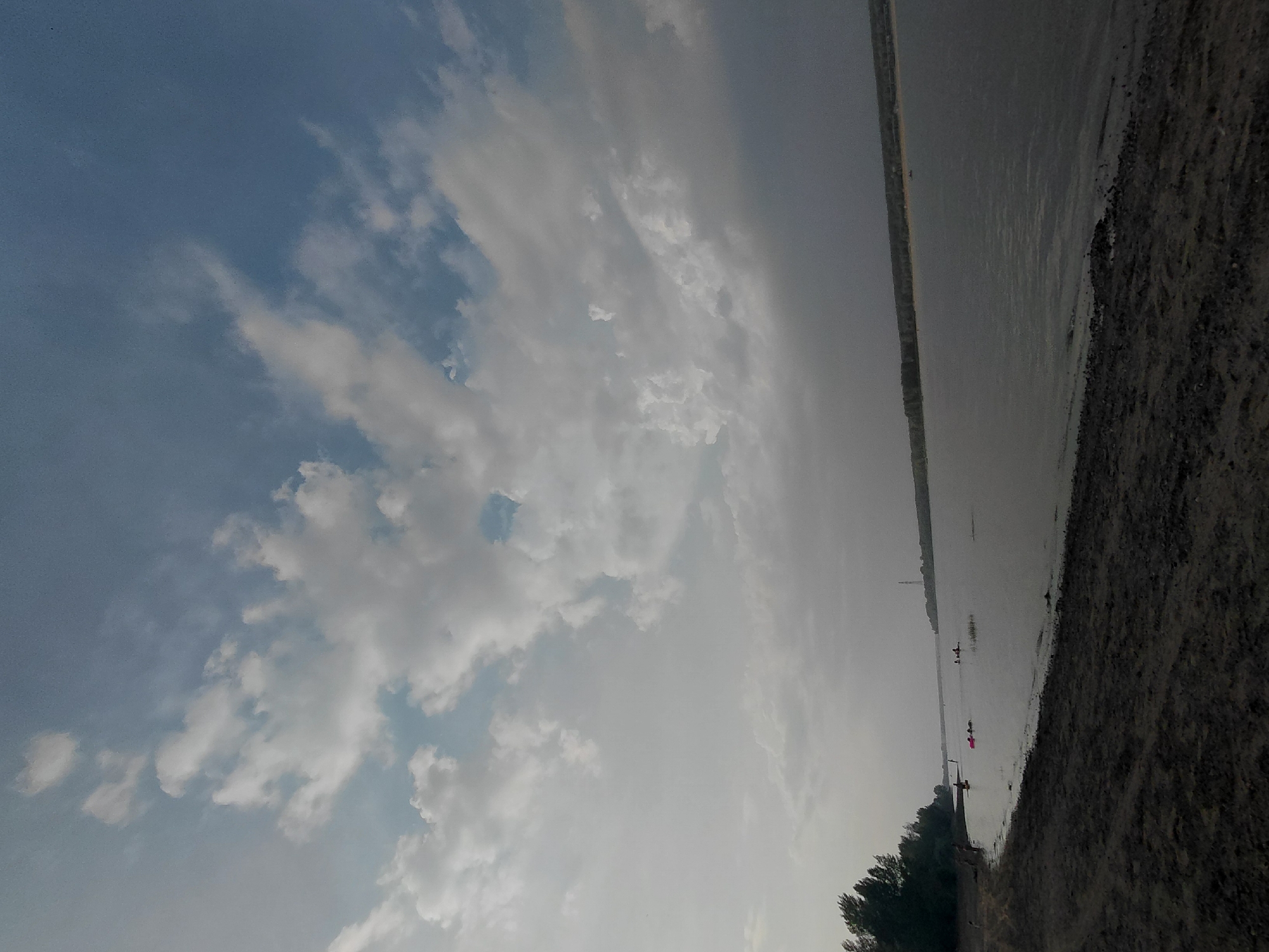 The image depicts a serene natural landscape, likely a riverbank, under a partly cloudy sky. The foreground features a sandy or muddy shore with some scattered debris, transitioning into a calm body of water. Several individuals are visible in the water, some appearing to be on kayaks or similar recreational watercraft. The distant bank of the river is lined with trees, with a faint structure, possibly a tower, visible in the far distance. The overall impression is one of a peaceful outdoor setting, perhaps a recreational area. The provided context suggests this scene is located in Novosibirsk, Russia, though the image itself does not contain explicit geographical markers.