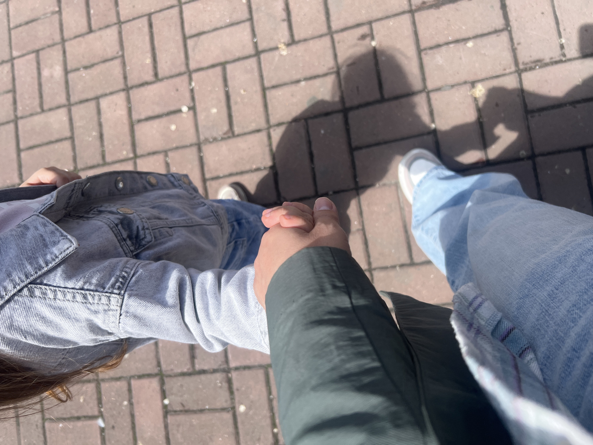 An overhead, full shot captures two people walking while holding hands on a paved surface in Khabarovsk, Russia. The image is taken from the perspective of one of the individuals, looking down at their interconnected hands and the path ahead.
The subjects include two individuals. On the left, a person is seen wearing a light-wash denim jacket and has brown hair visible near their shoulder. Their hand is intertwined with another hand, visible on the right. This second individual is wearing a dark green long-sleeved garment and light blue denim jeans. Their right foot, clad in a white sneaker with gray accents, is partially visible on the bottom right of the frame. Shadows of the two individuals are cast on the ground, indicating the presence of sunlight.
The activity taking place is two people walking together, holding hands. This suggests companionship or a romantic relationship.
Notable details include the time of day, which appears to be daytime due to the bright sunlight and distinct shadows. The weather seems clear and sunny. The location is an outdoor paved area, likely a sidewalk or pedestrian path, characterized by brick-like paving stones in varying shades of reddish-brown. Some light-colored debris or specks are scattered on the ground. There is no visible text in the image. The overall impression is one of a casual, pleasant walk.