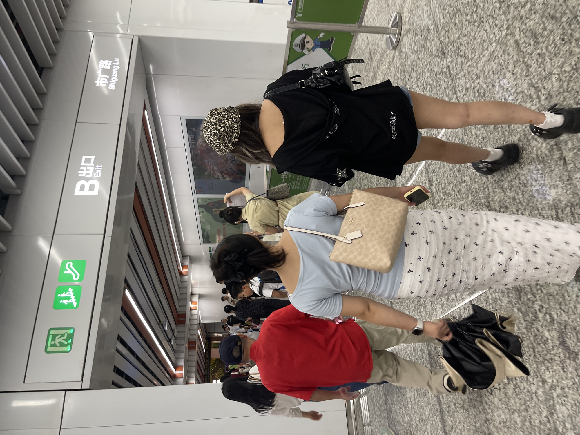 The image shows a group of people standing in a subway station. The station appears to be modern, with clean white walls and a speckled gray floor. Signs indicate "B Exit" and "Shiguang Lu" (Shiguang Road). There are also green emergency exit signs with icons depicting an escalator, a wheelchair accessible entrance, and a person exiting a door.
In the foreground, a person wearing a red shirt and khaki pants is holding a black umbrella. Next to them, a person in a light blue top and a white patterned skirt is holding a beige handbag. Further back, a person wearing a black top and a leopard-print hat is visible. Several other individuals are seen in the background, also appearing to be waiting or walking through the station.
The lighting suggests it could be daytime, but the indoor setting of a subway station makes it difficult to determine the exact time of day or the weather outside. The overall atmosphere seems to be one of routine transit, with people going about their day. No specific emotions are discernible from the image. The location is identified as Panyu District, China.