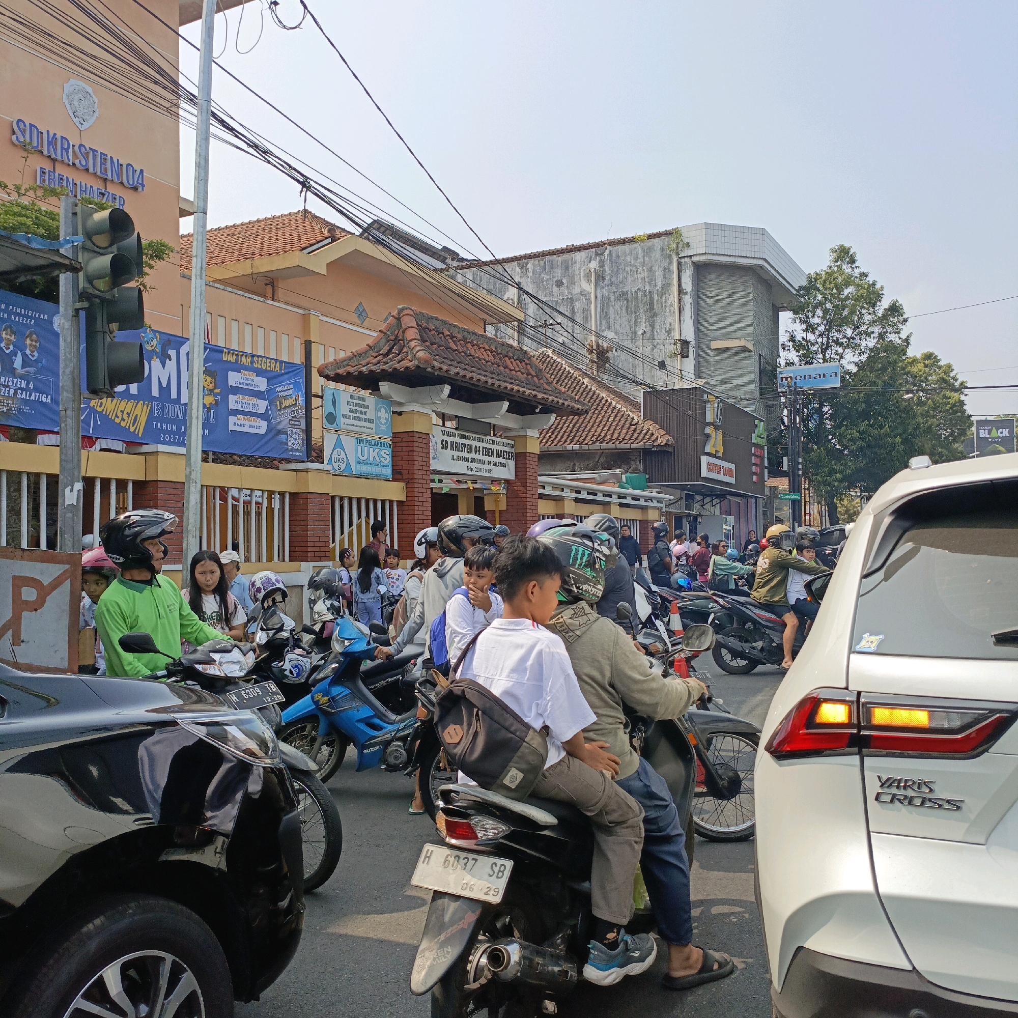 This image captures a street scene in Salatiga, Indonesia, likely near a school. The foreground is dominated by the rear of a white car, a black car, and numerous motorcycles with riders. In the mid-ground, a traffic light stands to the left, adjacent to a building with "SD KRISTEN 04 EBEN HAEZER SALATIGA" signage. Several blue banners with text and cartoon characters are displayed on the building's facade. People, including children and adults, are seen on and around the motorcycles, some wearing helmets. The environment is urban, with buildings, power lines, and a clear sky. The atmosphere appears to be a busy day, possibly during school drop-off or pick-up.
