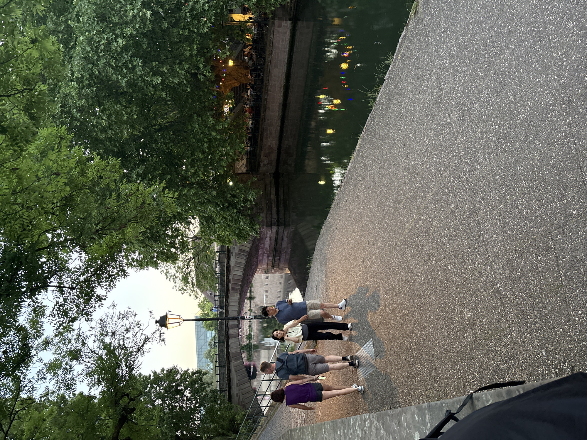 The image shows a group of four people standing on a paved path next to a canal or river. The path runs alongside the water, which reflects lights from buildings on the opposite bank. A stone bridge is visible in the background. The path is lined with trees. A lamppost stands near the group. The people appear to be posing for a photo or simply taking in the scenery. The location is likely in the city of Strasbourg, France.