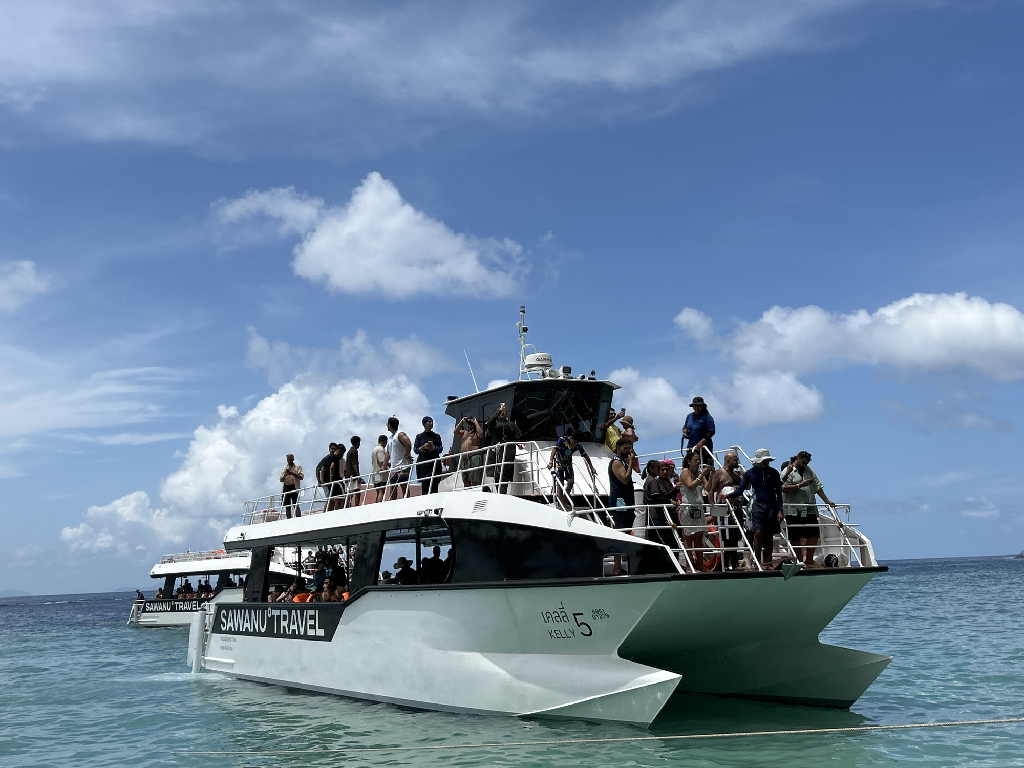 The image shows two white catamaran tour boats with many people on board, at sea with a clear blue sky and scattered white clouds. The boat in the foreground has "SAWANU TRAVEL" written on its side in black lettering, and also has the Thai script "เคลลี่ 5" and "KELLY 5" written on its hull along with a registration number. People are standing on the upper deck of the boat, some are looking out at the water, others are talking to each other. Some people are wearing life vests. The water is a clear turquoise color. The second boat, further in the background, also displays "SAWANU TRAVEL" on its side. The scene appears to be a scenic tour in the waters around Ko Lanta, Thailand.