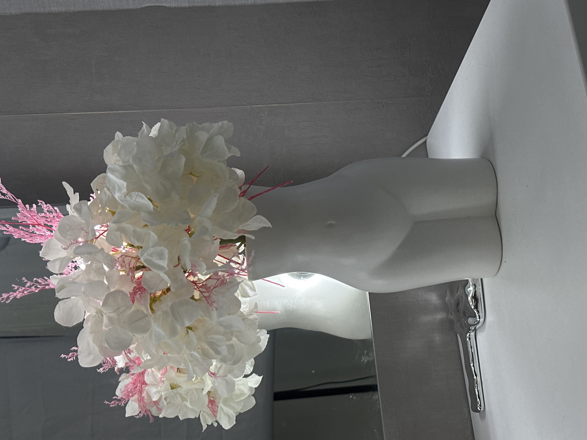 This image, captured in Sochi, Russia, is presented rotated 90 degrees clockwise. When viewed upright, it showcases an indoor decorative arrangement. The central focus is a white, torso-shaped vase, resembling a headless mannequin bust, serving as a container for a vibrant artificial floral display.
The bouquet consists primarily of large, lush white flowers, possibly hydrangeas, intermingled with delicate, thin pink decorative accents that resemble dried floral sprigs or fine plastic filaments. The vase rests on a smooth, white countertop or shelf that extends across the right side of the frame. Beneath the vase, a reflective surface, appearing to be a mirror or polished metal panel, captures and reflects a bright light source, likely a bulb, positioned to illuminate the scene from below. This creates a distinct glow and highlights the metallic elements.
In the background, to the left of the vase, is a textured grey wall. A thin white electrical cord is discreetly visible behind the vase against the white surface. Adjacent to the reflective panel on the right, there is a unique metallic, chrome-like decorative piece with a fluid, wavy design. The scene is a still life; no people are visible, and no specific activity is taking place. The overall aesthetic is modern and clean, characterized by a predominantly monochrome palette with a striking splash of pink. No text is visible within the image.