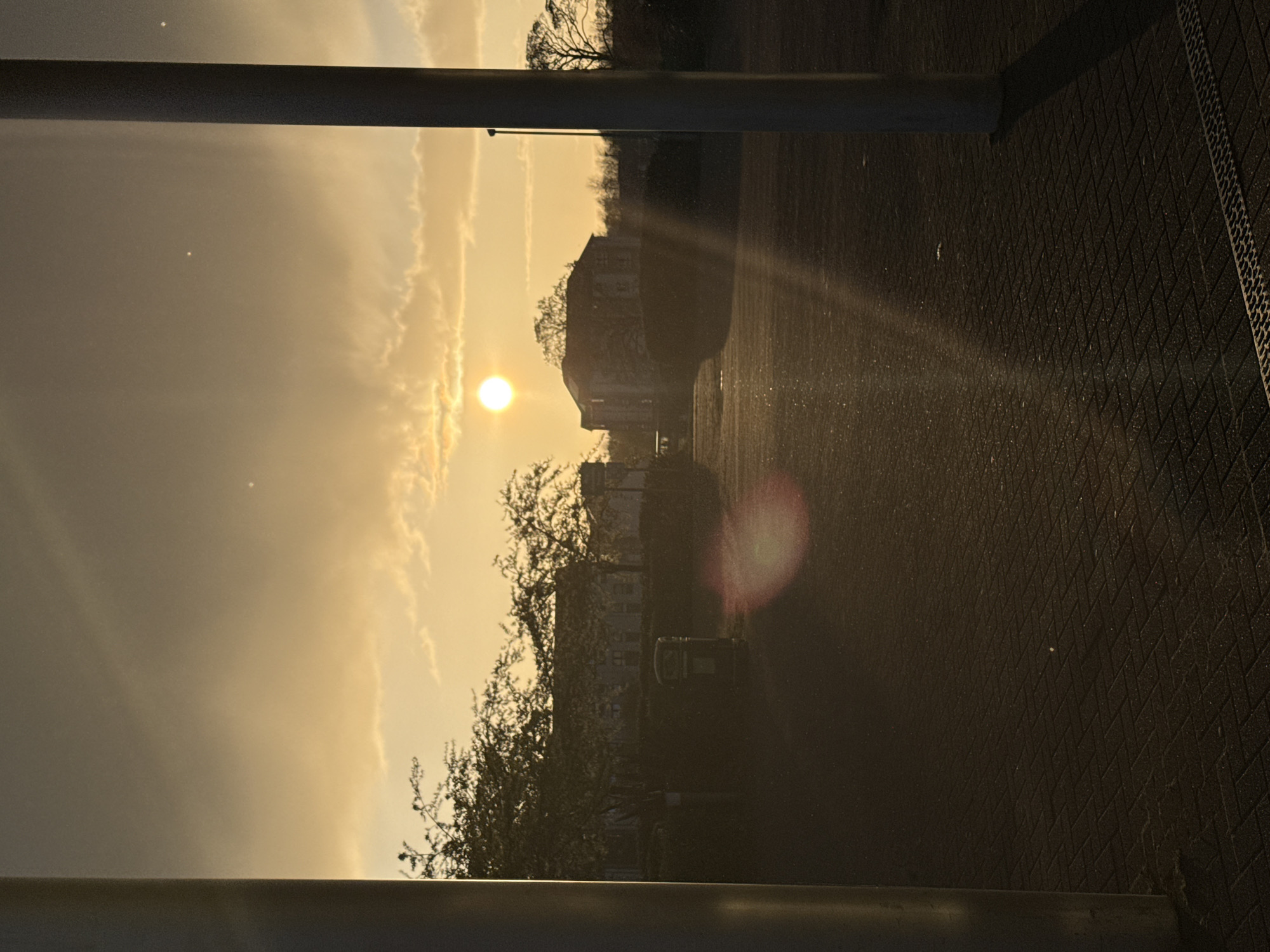 The image captures a sunset scene in an urban environment. The sun is low on the horizon, obscured by a building and trees. Two horizontal metal bars are located at the top and the bottom of the image, likely part of a structure like a fence or barrier. The ground appears to be paved with a pattern of small squares. There is a visible light beam emanating from the sun. The location is Unknown, Unknown.