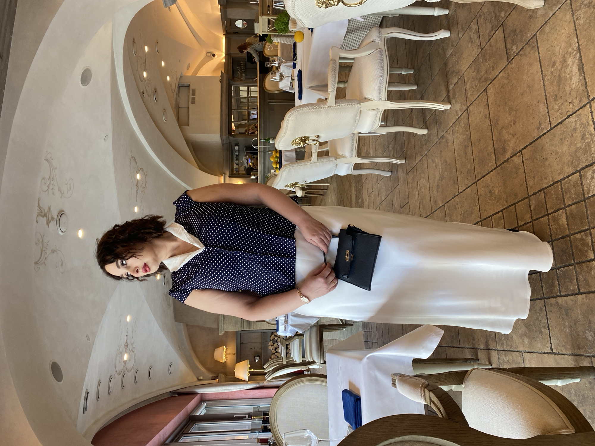 An elegant woman is captured posing within a sophisticated restaurant interior. She has dark, curly hair and is wearing a navy blue polka-dot blouse with a white collar, paired with a flowing, long white skirt. Her lips are adorned with red lipstick, and she wears a gold watch on her left wrist while holding a small black handbag or wallet in her hands.
The restaurant features an upscale ambiance with ornate white chairs, many having golden decorative elements, and tables draped in crisp white tablecloths. Some tables are set with wine glasses, dark blue napkins, and small decorative items like lemons or greenery. The floor is tiled with large, rectangular beige or brown tiles.
The ceiling is a prominent architectural feature, being arched and light-colored, adorned with intricate, light-toned scrollwork and multiple recessed light fixtures. These include circular spotlights and larger, rounder lights, casting a soft illumination. The walls also match the light color scheme, with some sections displaying a reddish-brown hue closer to the floor level. In the background, more dining areas, chairs, and what appears to be a counter or service area with staff are visible. The overall setting suggests a formal dining experience, possibly during the day with artificial lighting dominating, or in the evening. There is no discernible text visible in the image.