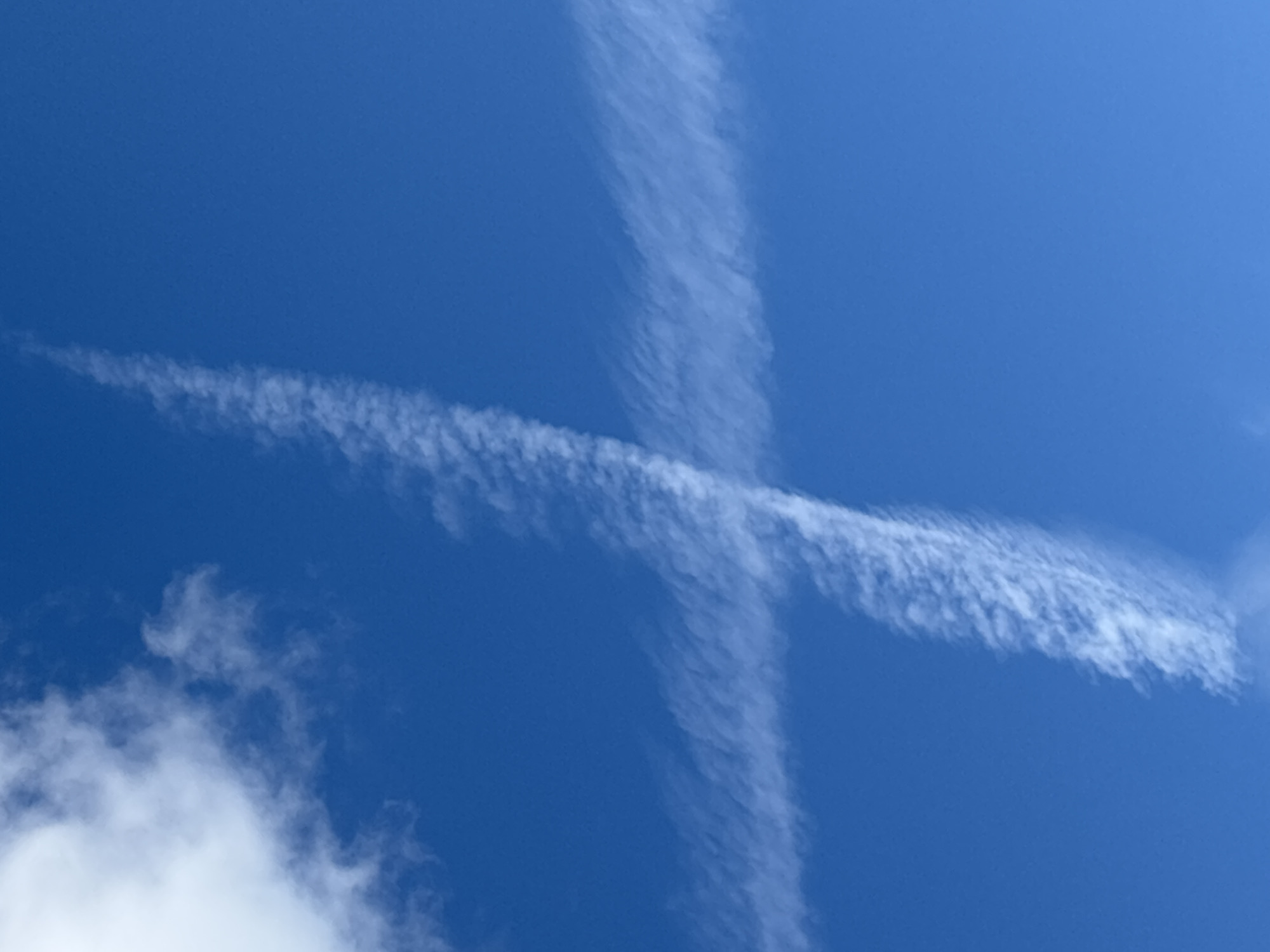The image displays a clear blue sky with two contrails crossing each other, forming an 'X' shape. One contrail runs diagonally from the bottom left to the top right of the frame, while the other runs vertically from the top to the bottom. The contrails are wispy and appear to be dissipating, suggesting they were formed by aircraft. In the bottom left corner, a small portion of a fluffy white cloud is visible. The scene is set outdoors, with no specific location cues other than the provided context of Vsevolodovo, Russia. The time of day appears to be daytime, given the bright blue sky. There is no activity or event taking place, beyond the natural formation and dissipation of contrails. No people, objects, or text are visible in the image.
