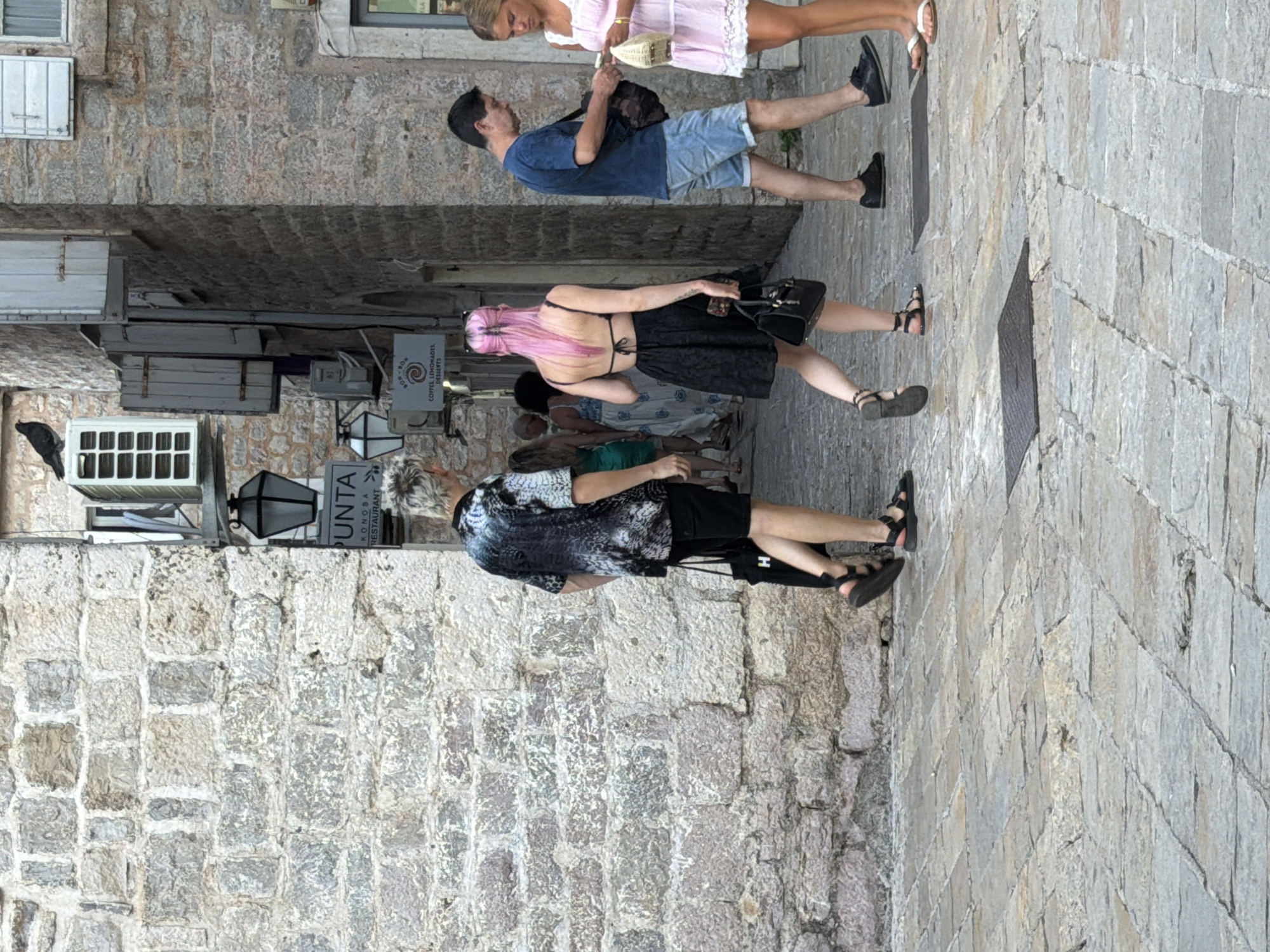 In Budva, Montenegro, a group of people walk along a narrow cobblestone street. On the left, a man with a distinctive black and white patterned shirt and black shorts walks away from the camera, carrying a black tote bag. Beside him, a woman with bright pink hair and a black dress with a low-back design walks ahead, also carrying a black handbag. Further ahead, a man in a blue t-shirt and denim shorts walks, holding a bag, while a young woman in a pink lace dress stands to his right. The street is lined with old stone buildings, one of which features a sign for "PUNTA KONGOBA RESTAURANT." The architecture suggests a historic urban setting.