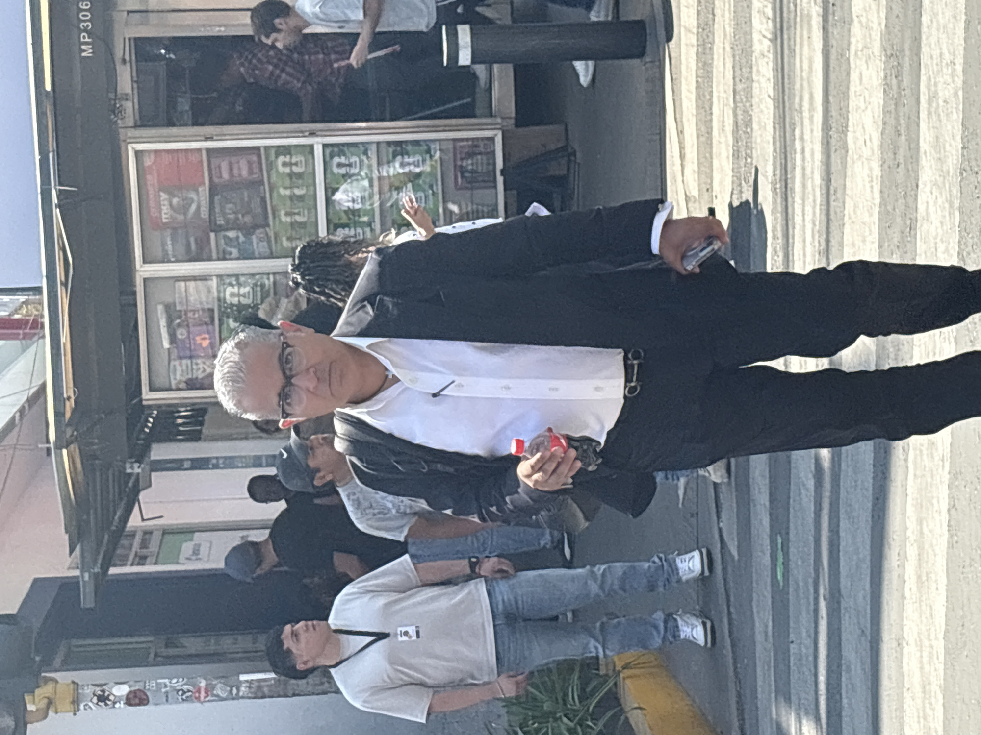 This image depicts a bustling outdoor urban street scene during daylight hours, likely mid-day given the bright lighting and distinct shadows. The central figure is a man with grey hair and glasses, wearing a black suit jacket, a white collared shirt, and dark trousers. He holds a small red bottle in his left hand and a mobile phone in his right, looking directly at the camera with a calm expression.
Behind him and to his left, a storefront or stall is visible with a display window containing various items, possibly magazines or comics. Visible text on these items includes "STREET", "TRAY", and "COMICS". Inside this establishment, a young man in a plaid shirt appears to be playing a percussion instrument with drumsticks. Above him, another person in a white t-shirt is partially visible.
Several other people are in the background, mostly obscured. To the main subject's left, a man in a light t-shirt and blue jeans wears an ID badge on a lanyard. Further back, a person with dreadlocks is partly visible. The ground is paved, showing vertical stripes from the sun's reflection or a textured surface, and a yellow curb or bollard is visible in the lower right. A dark, horizontal structure with the text "MP306" is present in the upper left, potentially a part of a street sign or building canopy. The setting suggests a lively city street, consistent with Mexico City.