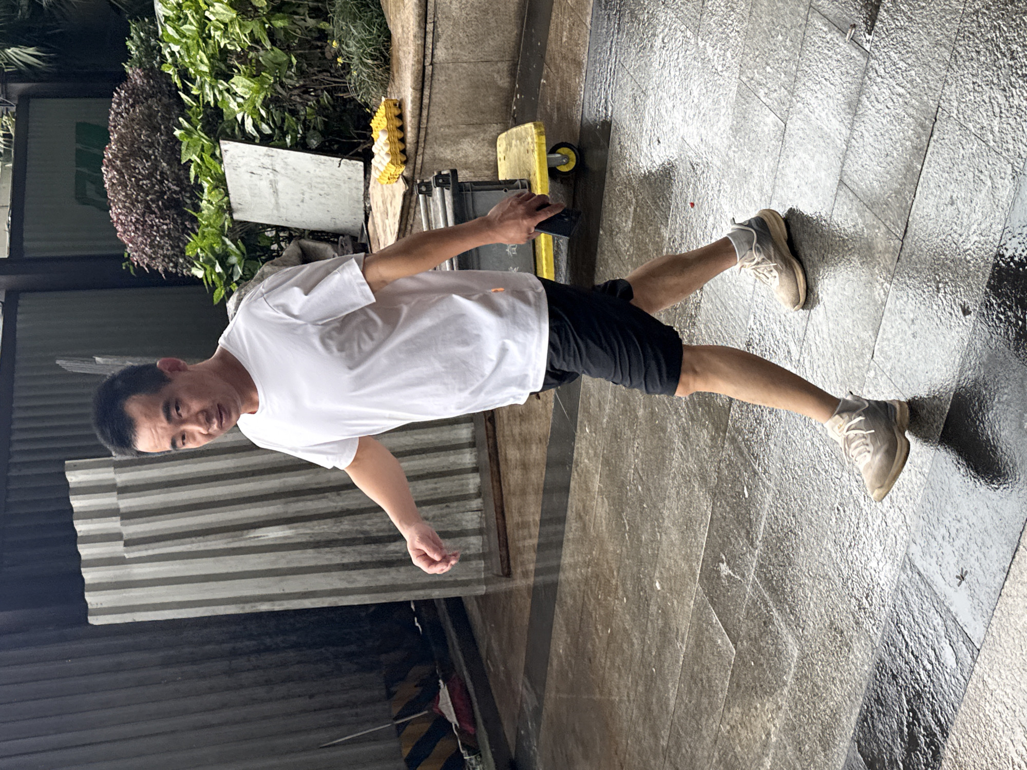 A man stands on a wet, gray tiled ground in an urban outdoor location. He wears a white t-shirt, black shorts, and tan sneakers. He appears to be holding a black object in his right hand. Behind him are plants, a white sign, a yellow dolly, and metal fencing. The scene is located in Nan'an District, China.