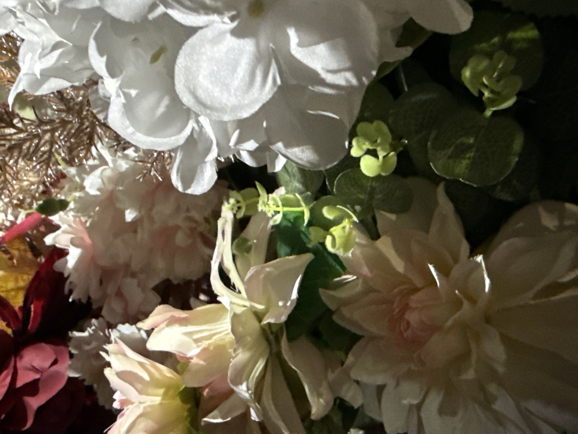 The image shows a close-up of an arrangement of artificial flowers. The flowers are various colors including white, pink, red and yellow. There are also green leaves and some brown decorative pieces. The light is dim. The location, Tyulyuk, Russia, is not discernible from the image.