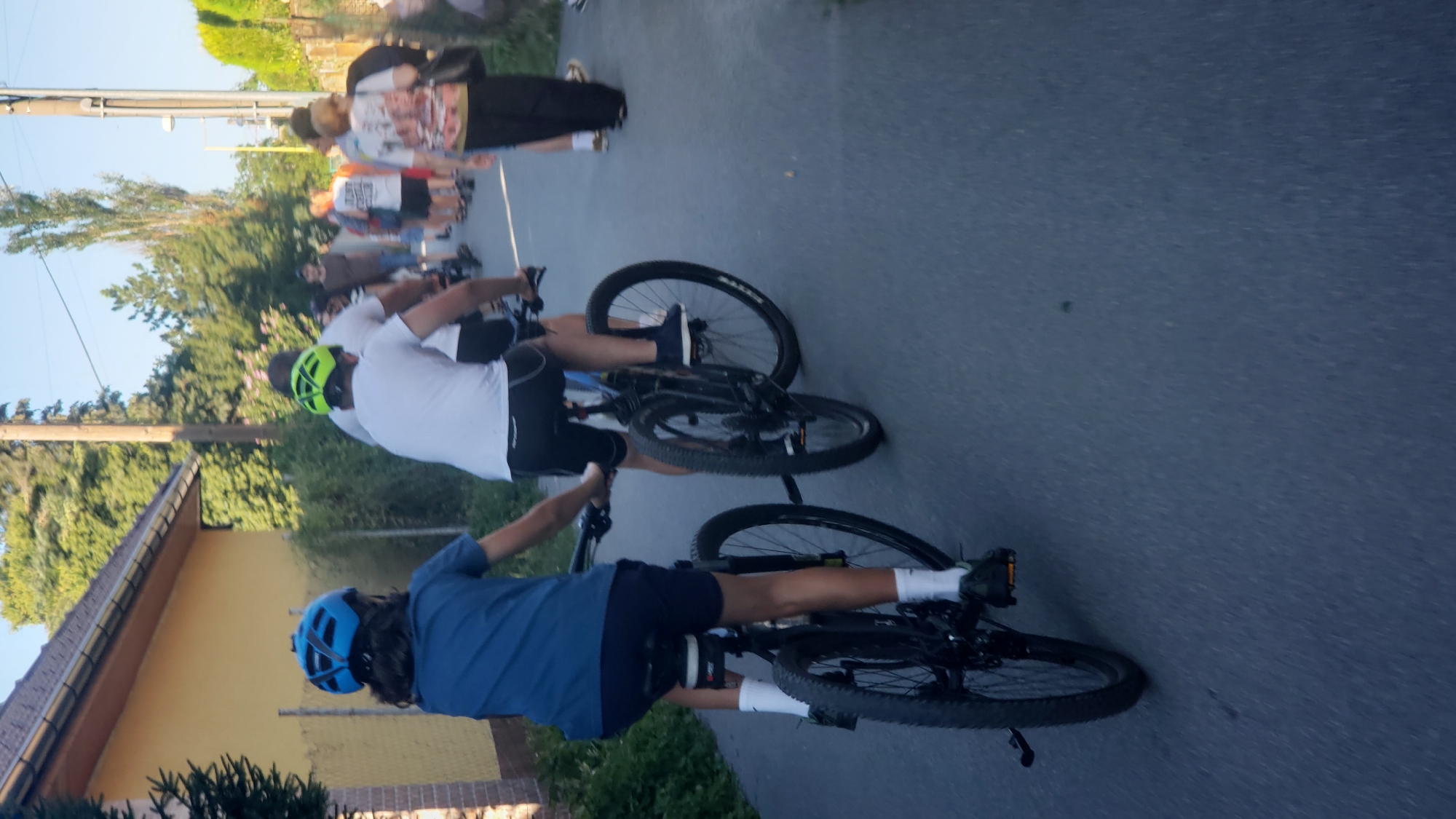 A group of people are cycling and walking down a paved street in Prešov, Slovakia. In the foreground, two children on bicycles are moving away from the camera. The child on the left wears a blue t-shirt and dark shorts, with a blue helmet. The child on the right wears a white t-shirt and dark shorts, with a neon green helmet. Behind them, other people are walking and cycling. The street is lined with trees and buildings, one of which is a yellow house with a brown roof. The sky is clear and blue.