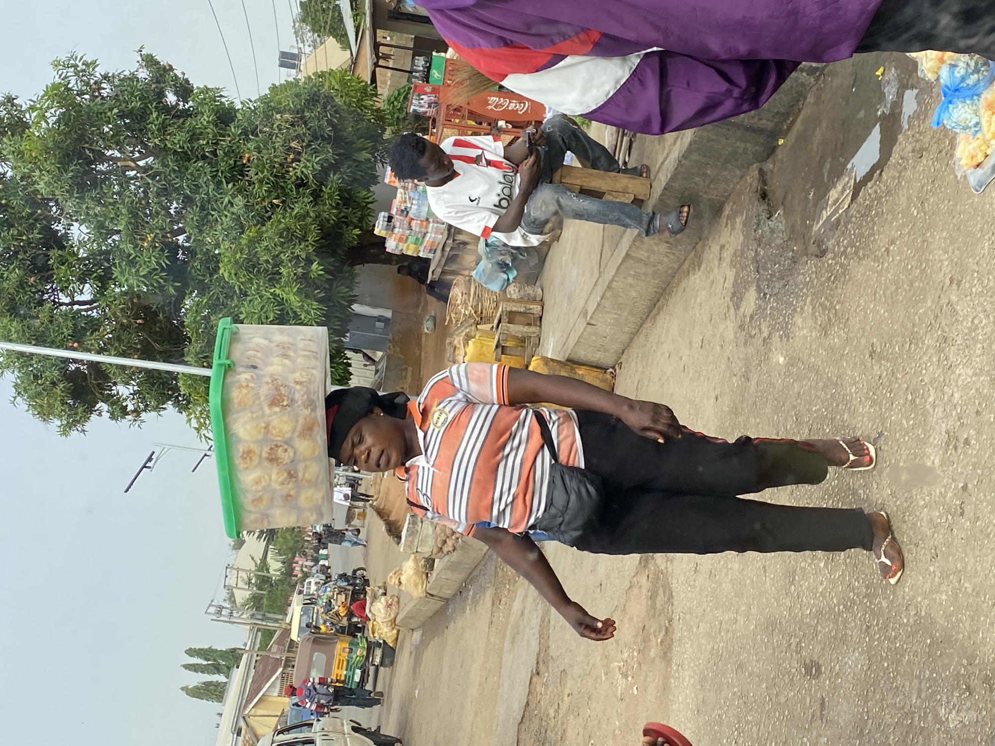 A vendor is carrying a container of baked goods on her head in Bukuru, Nigeria. The container is green and clear, revealing rows of baked goods. The vendor wears a striped orange and white shirt with black pants, a black head covering, and flip-flops. She carries a black bag across her body. The background shows a busy street scene with pedestrians, motorbikes, cars, and three-wheeled vehicles. A tree is on the left side of the image. Other vendors and pedestrians are on the side of the street. A man sits on a wooden crate nearby, using a mobile phone. A Coca-Cola sign is visible on a nearby stall.