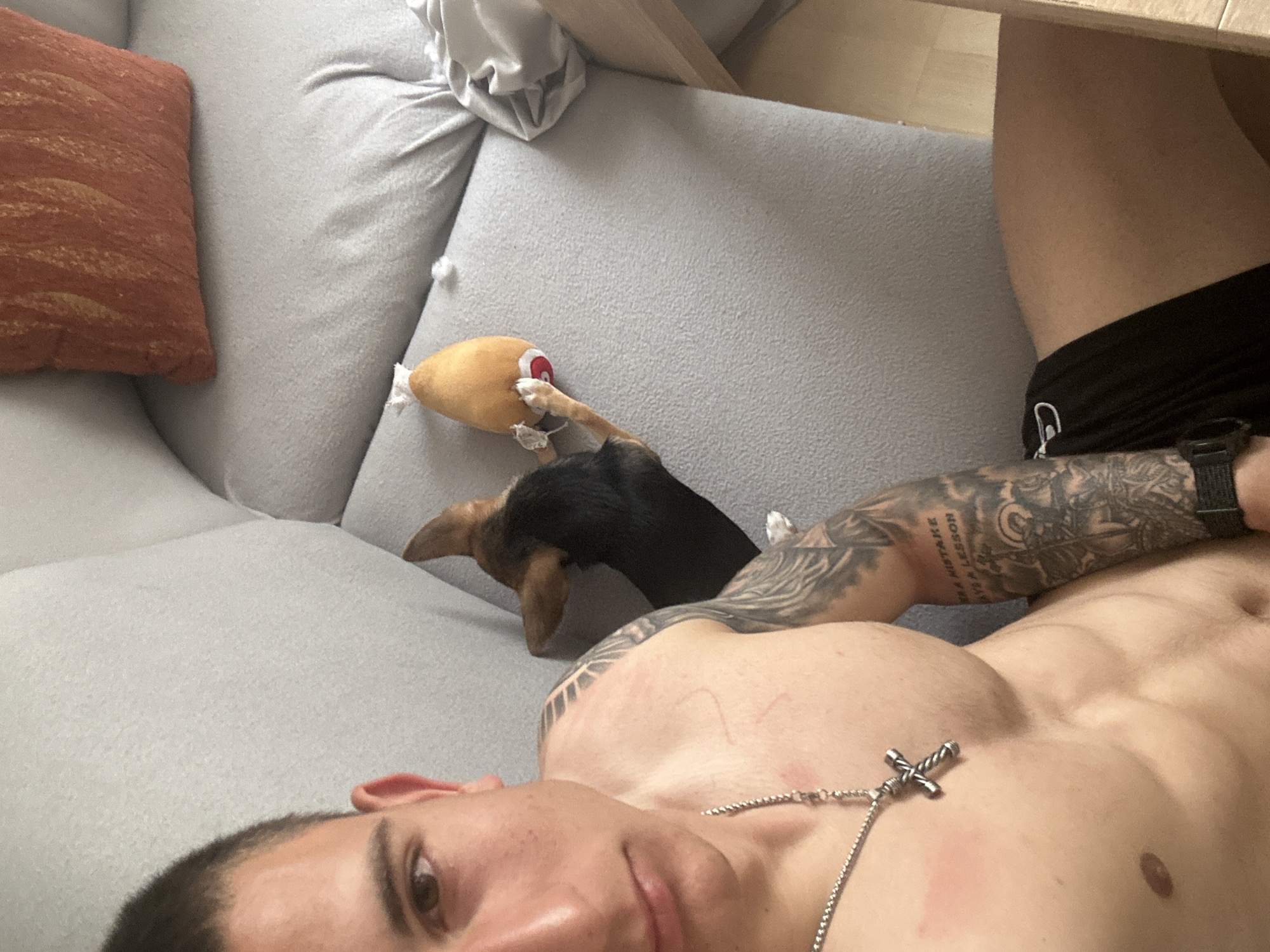 A shirtless man with a tattoo on his left arm, wearing a silver cross necklace, is lying on a light gray couch. A tan and black dog is lying next to him, with its paws on a yellow plush toy. An orange pillow is visible on the left. The location is likely an indoor living space. There is no information to determine if this is in the city of Burgas, Bulgaria.