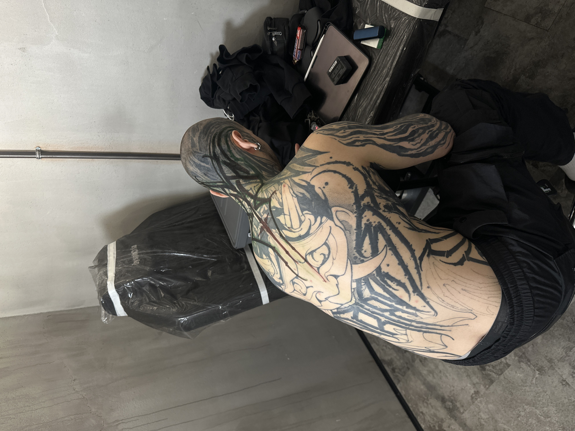 The image depicts a person, appearing male, seated in what looks like a tattoo studio or workshop. The person is viewed from the back, with their head slightly turned, revealing extensive blackwork tattoos covering their entire back, neck, and arms. Their hair is shaved on the sides with the top slicked back, also featuring black tattoos. A silver ring earring is visible in the left ear.
The back tattoo is a large, intricate Japanese-style design, possibly an oni or Hannya mask, with detailed linework. Some red marks are visible on the back, suggesting fresh tattooing or blood. The arm tattoos feature abstract or tiger-stripe-like patterns, with some areas still in outline form.
To the person's right, on a dark table or surface, are various items: a neatly folded black hoodie, a black tactical bag or vest, a tablet, a black electronic device (possibly a power bank), a white stylus, a blue pen case, and a "Snickers" candy bar. To the person's left is a black, rectangular object, possibly a padded bench or chair, entirely covered in clear plastic wrap. A white strap or tape is visible across it, along with the partial text "Dysansia."
The background consists of light grey, textured concrete walls, with a horizontal metal pipe and a clamp running across one wall. The floor is also grey, resembling concrete or industrial tiling. The scene suggests an indoor setting, likely in Moscow, Russia, where the person is either undergoing a tattoo session or has recently completed one.