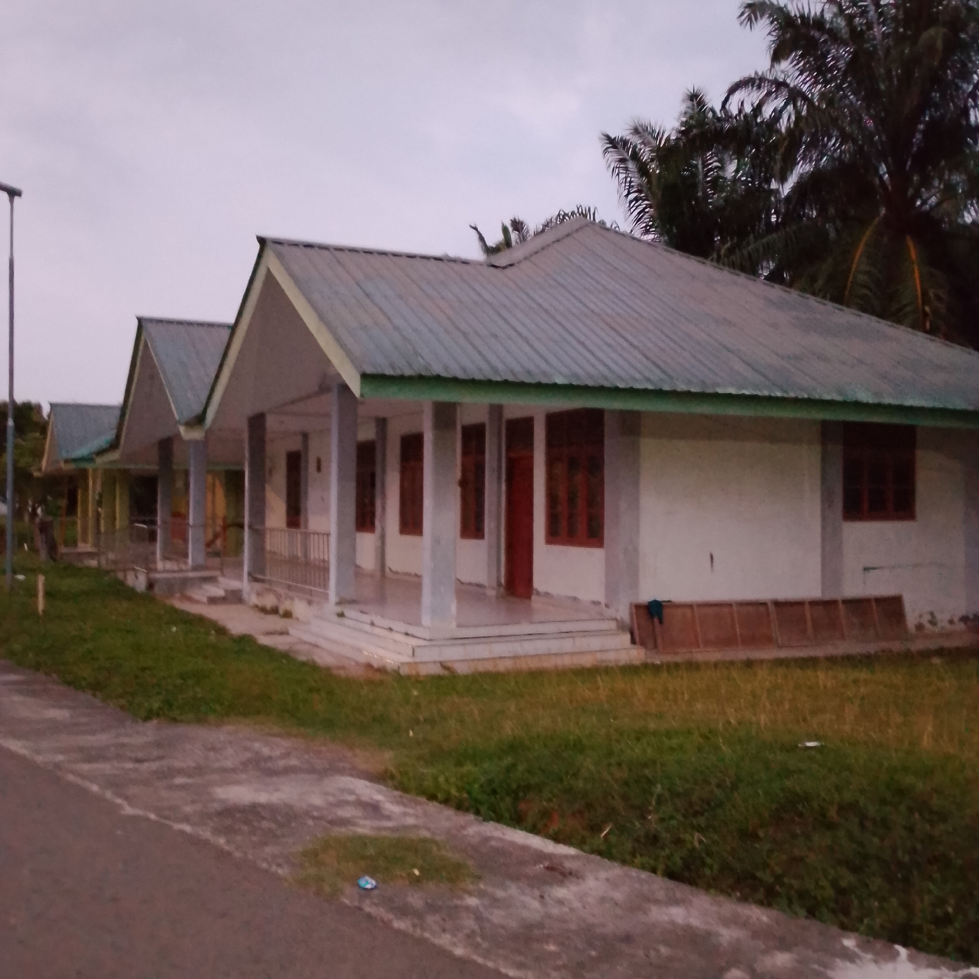 The image shows a row of identical, small, single-story houses with peaked, metal roofs. The houses are painted white and have light-colored porches with pillars. There are multiple windows with dark wooden frames on each house, and at least one visible red door. In front of the houses, there is a paved walkway and a grassy area. In the background, palm trees are visible. The sky is overcast and the lighting suggests it is either dawn or dusk. The scene appears to be in a tropical or subtropical climate. There are no people visible in the image.