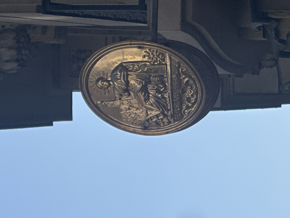 An oval-shaped, dark gold or bronze metallic relief medallion, bearing the inscription "RÉPUBLIQUE FRANÇAISE," is affixed perpendicularly to the light-colored stone facade of a building. The medallion features a seated allegorical female figure, likely Liberty or Marianne, wearing a radiated crown. She holds a fasces in her left hand and a rudder in her right, with wheat sheaves and other foliage visible beside her. Architectural details of the building, including cornices and moldings, are present to the right and above the medallion. A clear, light blue sky occupies the upper left portion of the frame. The medallion is illuminated by direct sunlight. This emblem indicates the location as a public or governmental building within the city of Paris, France.
