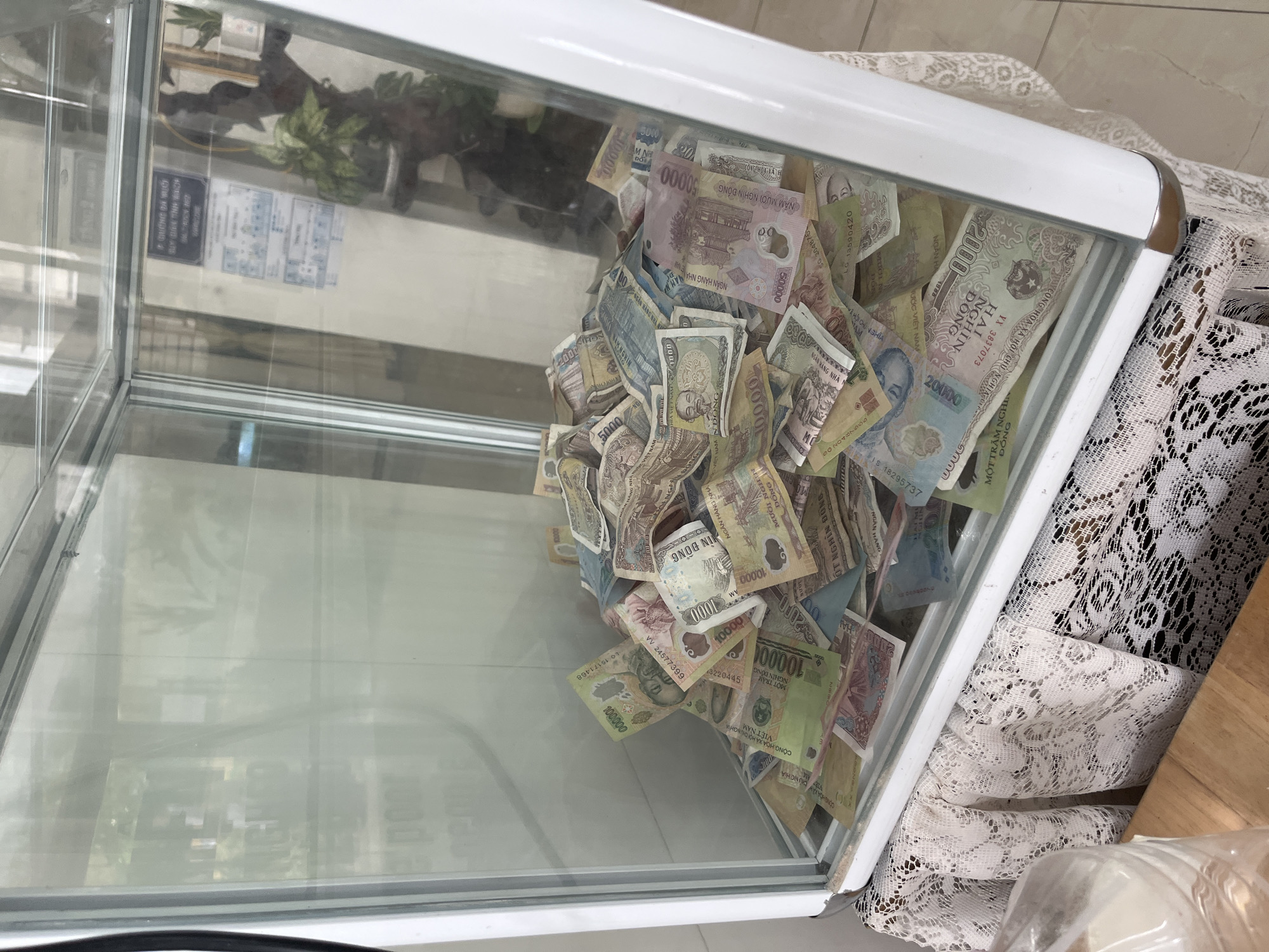 A clear glass donation box with a white frame is filled with a dense pile of Vietnamese Dong currency. The box sits on a surface covered with white lace. A partial view of a wooden table is visible to the right. Reflections in the glass show an out-of-focus interior and signage. The cash includes denominations such as 20,000, 10,000, 50,000, 2,000, 100,000, 1,000, and 5,000 Dong. The printed text identifies some of the banknotes as being issued by the State Bank of Vietnam (Ngan Hang Nha Nuoc Viet Nam). The location is indicated on one note as being in Nam Nha Trang Ward, Vietnam.