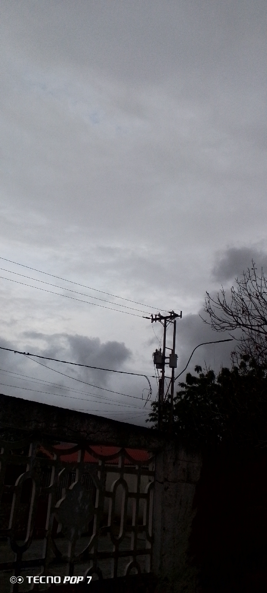 The image depicts a gloomy sky with heavy clouds, suggesting an overcast or stormy day. In the foreground, a dark, ornate fence or gate is visible, with decorative metalwork. Above the fence, utility lines stretch across the frame, supported by a wooden utility pole with two transformers mounted on it. To the right of the pole, a bare tree branch extends into the sky, and dark foliage is visible in the lower right portion of the image. The overall scene is one of a residential or urban periphery, with a focus on the infrastructure against a somber sky. The bottom left corner of the image contains a watermark that reads "TECNO POP 7".