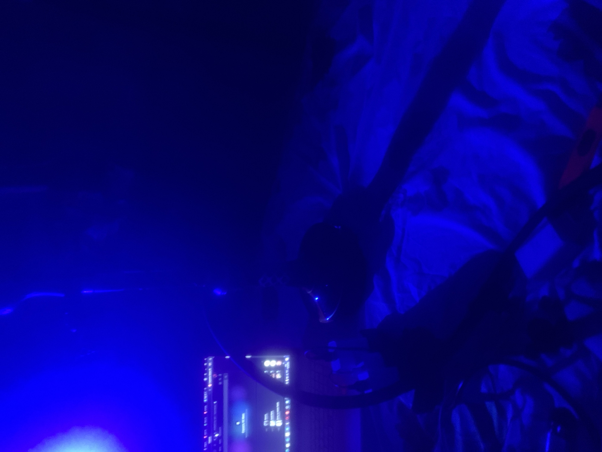 The image captures a dimly lit indoor setting, predominantly bathed in a deep blue artificial light. The perspective is slightly askew, suggesting a personal space like a bedroom or leisure area during nighttime.
In the lower left quadrant, a computer monitor displays a graphical user interface. The screen, glowing with blue and white light, shows what appears to be a desktop environment with various icons and windows, though no specific text is legible.
Towards the center of the frame, a shadowy figure of a person is partially visible, wearing a pair of headphones or a headset that subtly reflects the blue light. The person's arm or hand can be seen on the right, resting on a textured surface, which appears to be rumpled bedding or blankets.
On the far right, near the bedding, a distinct reddish-orange object with a black circular component is visible. This object resembles a gaming controller or a part of a virtual reality system. Other dark, indistinct shapes and lines throughout the image may represent cables, furniture, or other equipment.
The scene conveys an atmospheric, low-light environment, characteristic of someone engaging with a computer, likely gaming, working, or consuming media, late in the evening or at night. No specific location outside of an indoor private room is identifiable.