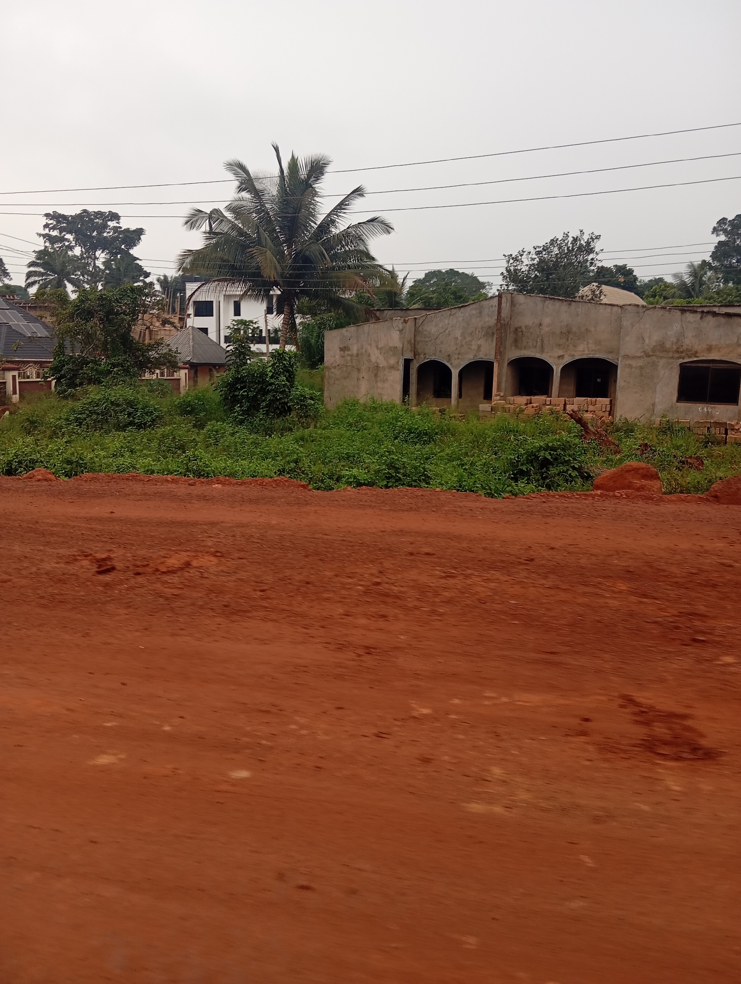 A construction site is depicted in Nsukka, Nigeria, under an overcast sky. In the foreground, a dirt road with a reddish-brown hue is visible. Behind the road, lush green vegetation covers the ground, with some piles of red earth and bricks scattered amongst it. Further back, a partially constructed building stands with arched doorways and windows. To the left of this building, a modern white house with dark windows is visible, surrounded by trees, including a prominent palm tree. Electrical wires are strung across the top of the image, against the pale sky. The overall scene suggests ongoing development in a semi-rural or suburban area.