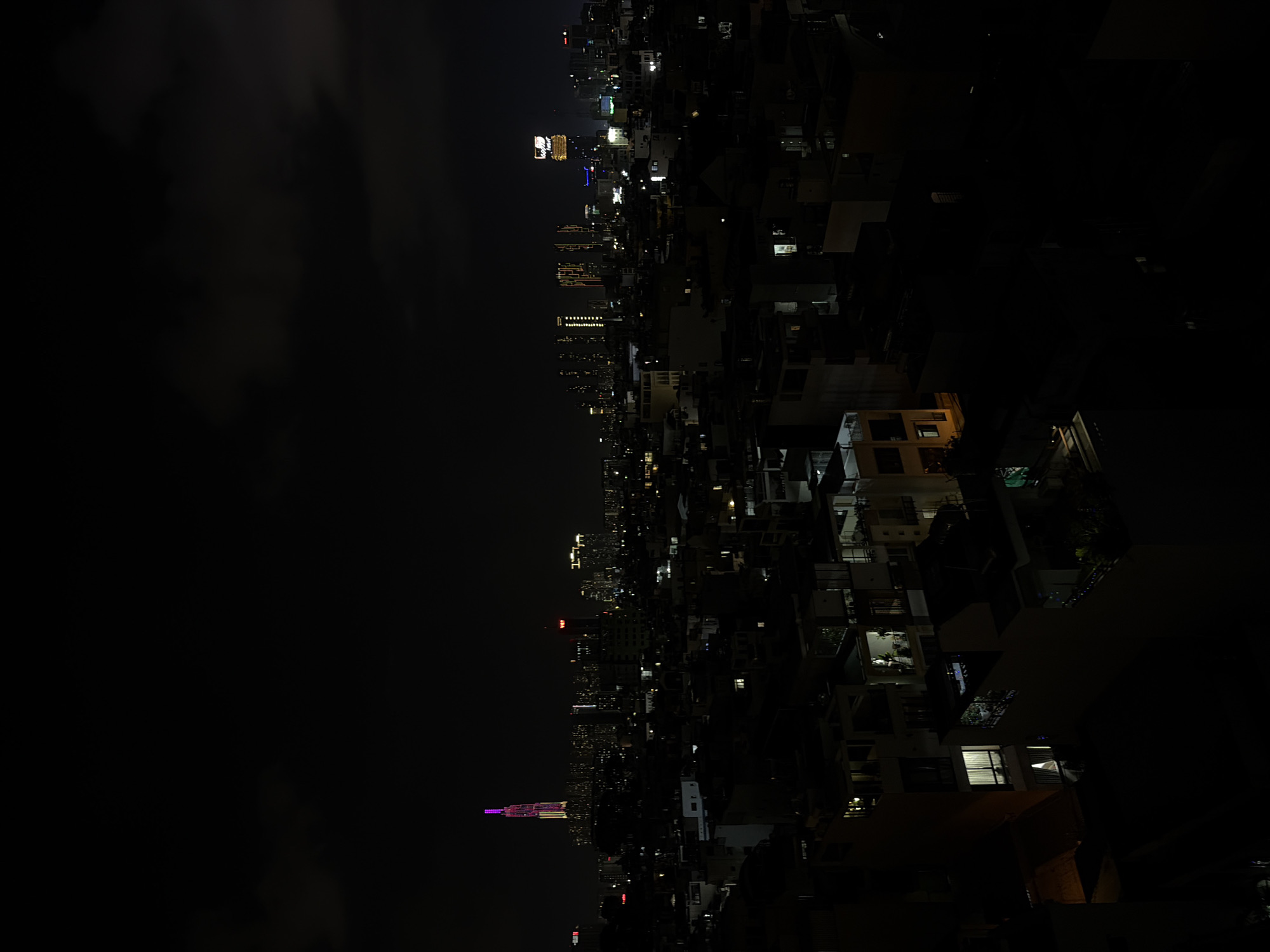 The image captures a panoramic night view of a dense urban landscape, likely Thủ Đức, Vietnam, observed from an elevated perspective. The left half of the frame shows a vast expanse of dark night sky with subtle cloud formations and atmospheric haze.
The right side of the image reveals a sprawling cityscape, characterized by numerous buildings of varying heights, including residential and commercial structures. These buildings are illuminated by a multitude of artificial lights, with windows emitting warm yellow and bright white glows against their dark facades.
A prominent landmark in the mid-distance is a tall, slender skyscraper, distinctively illuminated with purple and white lights on its upper sections. This architectural feature strongly suggests it is Vincom Landmark 81, a notable building in Ho Chi Minh City. Along the distant horizon, other high-rise buildings contribute to the city's extensive glow.
In the closer right foreground and mid-ground, the rooftops and upper levels of various buildings are visible, showing individual lit rooms and some architectural details. A large, brightly lit sign is discernible on a building in the middle distance; although the text is not clearly legible due to resolution, it appears to be a corporate name, possibly "VINCOM," as indicated by closer inspection in hint images. The overall scene conveys the vibrant and expansive nature of a city at night.