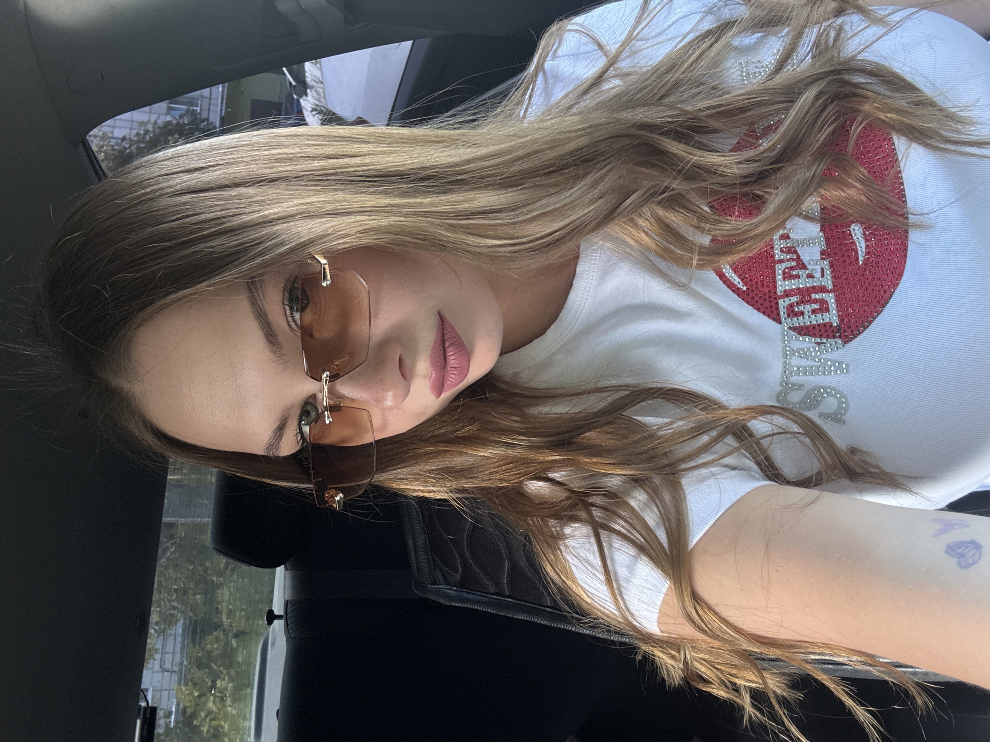 A young woman with long blonde hair, wearing sunglasses and a white t-shirt with a sequined lip design, poses for a selfie. She is sitting in the passenger seat of a car, with the window reflecting trees and buildings outside. The photo appears to have been taken during the day, with bright sunlight illuminating the scene. The woman's expression is neutral, and her makeup is subtle. A tattoo of a butterfly is visible on her arm. The text "BFF" is visible on her t-shirt, in rhinestones.