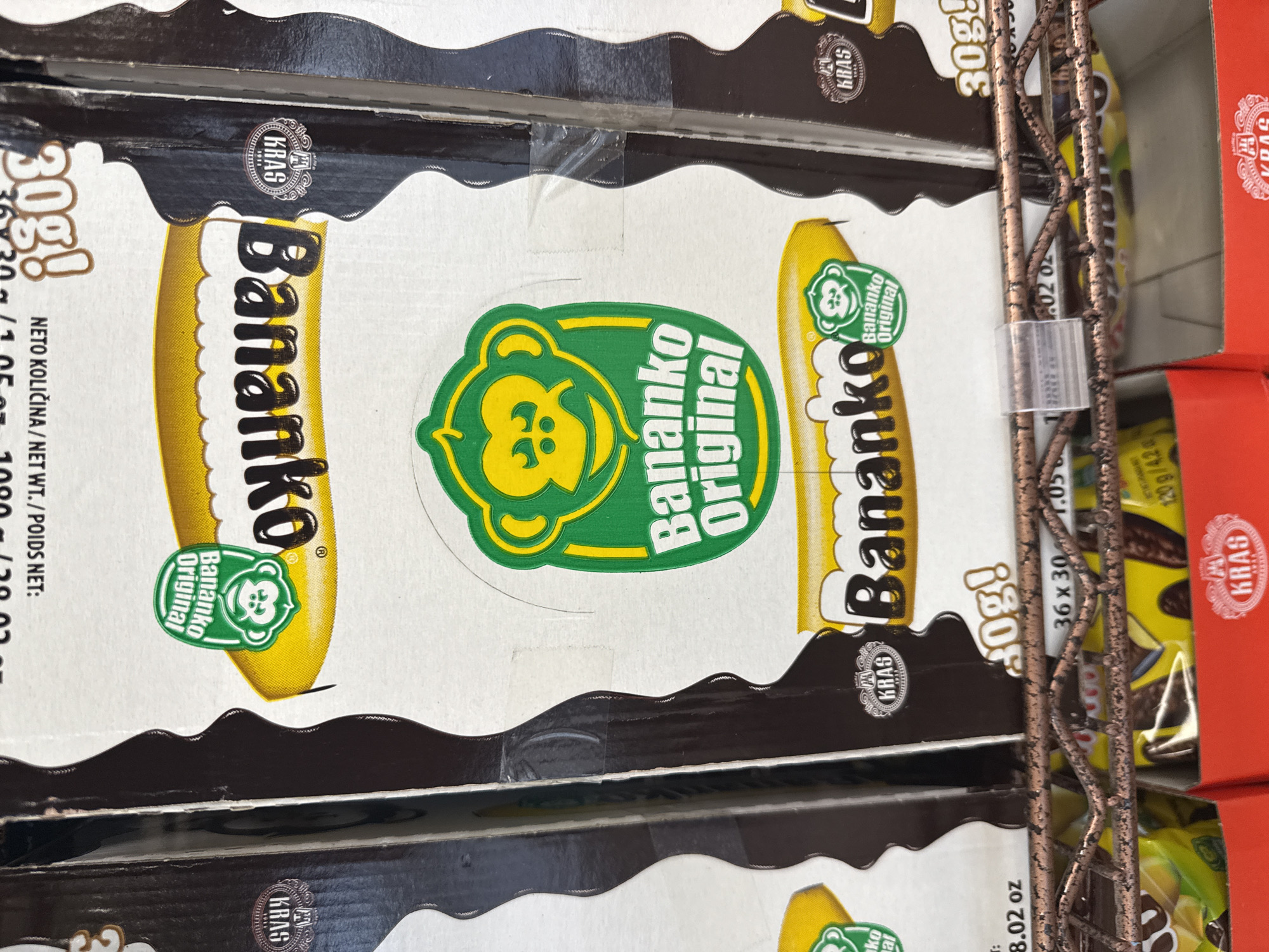 The image shows stacked boxes of "Bananko Original" chocolate-covered banana candies on a metal wire shelf, likely in a retail environment. The packaging is white with a yellow banana graphic, the brand name "Bananko" in black letters, and a green logo with a monkey. The boxes have brown tops and scalloped edges. The brand "KRAS" is also visible on the boxes. Text on the boxes indicates the product weight "30g," and "NETO KOLIČINA/NET WT./POIDS NET: 1080g/38.02 oz." The shelf is likely in a grocery store, possibly in Darien, USA. A red box is seen to the right of the Bananko boxes.