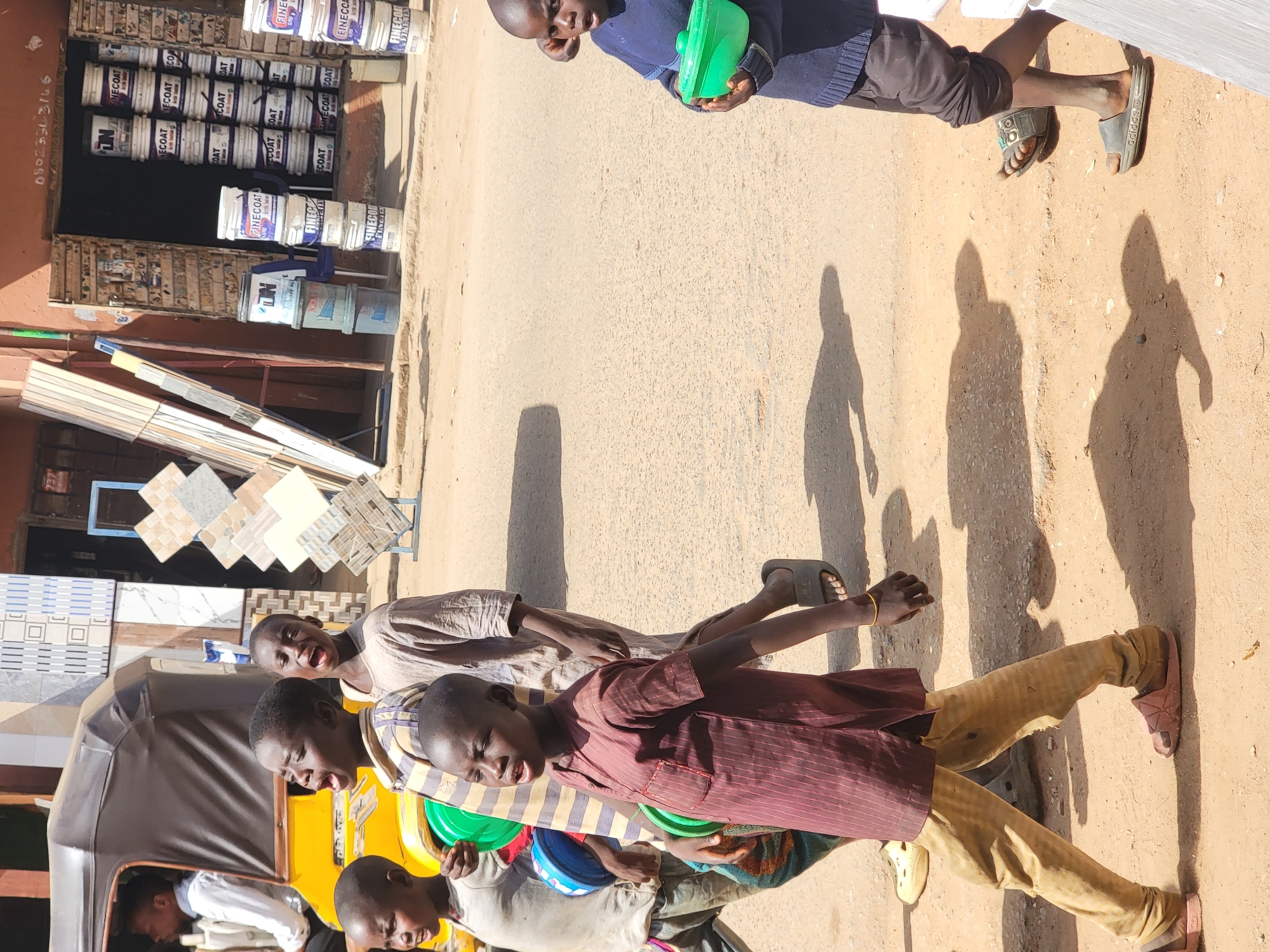 The image, apparently taken in Maiburiji, Nigeria, shows a group of boys situated alongside a roadside shop. The shop is lined with paint cans stacked in rows, and also features tiles on display. Several boys are gathered together, with some carrying what appear to be green and blue buckets. One boy stands apart, further up the road, also holding a green container. The ground is light-colored, and shadows of the boys stretch across the road surface. An auto rickshaw is partially visible, parked close to the shop.