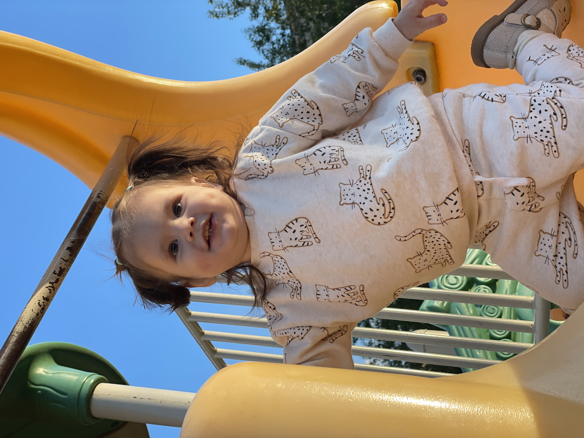 A toddler is on a yellow slide at a playground. The child is wearing a light-colored, cat-printed sweatshirt and matching pants. The child is smiling and looking at the camera. The playground equipment is visible in the background. The location in Izhevsk, Russia cannot be determined from the image.