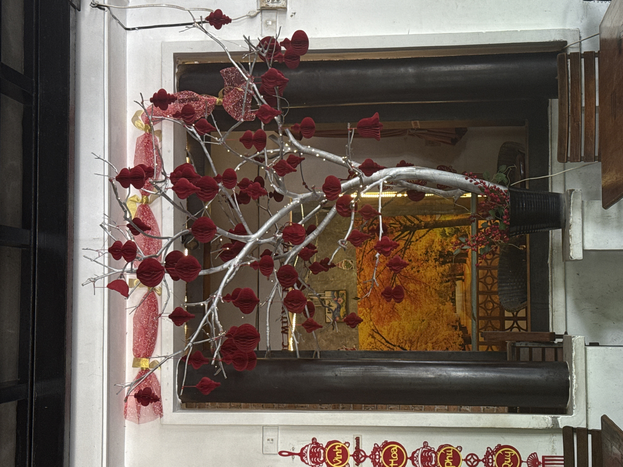 The image depicts a festive display, likely for Tet (Vietnamese Lunar New Year), in Nha Trang Ward, Vietnam. At the center is a silver-colored artificial tree adorned with numerous red, honeycomb-shaped ornaments. The tree branches are also decorated with small, white fairy lights. Behind the tree, a doorway reveals a view of a colorful tapestry or mural featuring trees with golden foliage and a pathway, suggesting an autumn or celebratory theme.
To the left of the tree, a vertical string of decorative red and yellow ornaments hangs on a white wall. These ornaments appear to be traditional Vietnamese designs, with figures and circular medallions bearing text in Vietnamese, such as "Vinh," "Hoa," "Phú," and "Qúy."
In the foreground, a dark-colored planter with red berries and green leaves sits on a ledge. The scene is set outdoors, possibly on a patio or verandah, as evidenced by wooden chairs and tables visible at the bottom of the frame. The architectural elements include white walls, dark pillars, and a dark ceiling structure at the top. The overall atmosphere is one of vibrant decoration and cultural celebration.