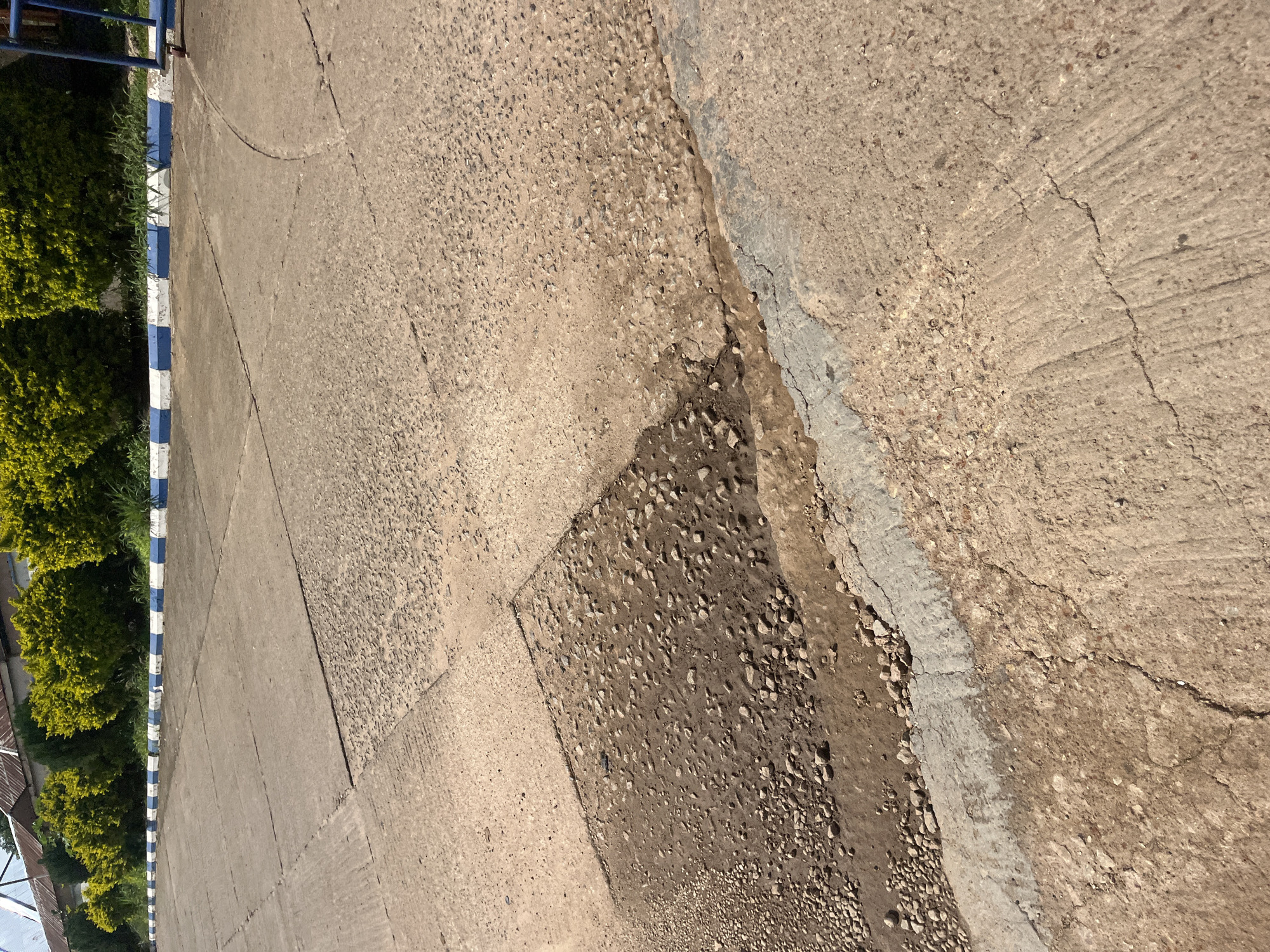 The image depicts an exterior scene of a paved area, possibly a parking lot or roadway, in Bukuru, Nigeria. The pavement is made of concrete slabs and shows signs of wear and tear, including cracks and patches of exposed aggregate and dirt. A section of the pavement in the foreground has a significant depression filled with soil and gravel, indicating a localized area of damage or erosion. A light-colored line, possibly a painted curb or edge marking, runs along the bottom edge of this damaged section. In the background, a row of green, well-maintained bushes or hedges is visible, with a building or roofline behind them. A blue and white striped border runs along the edge of the paved area, adjacent to the greenery. The overall impression is of a somewhat neglected but functional outdoor space.