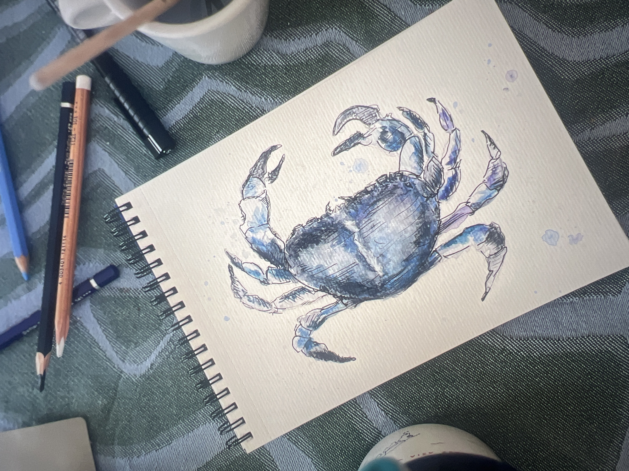 An overhead photograph captures an open spiral-bound sketchpad displaying an artwork on a textured, off-white page. The drawing depicts a blue and purple crab, rendered with watercolor washes and black ink outlines, situated diagonally across the page. Small blue paint splatters are visible around the crab. Surrounding the sketchpad are several art supplies: a light blue colored pencil, a dark blue colored pencil, a black pencil, an orange-brown pencil with "4 003401 121114" and "12111-101" visible on its barrel, and a black capped marker or pen. A partially visible white container holding other wooden instruments is located in the upper right. A cylindrical object with teal and white elements is visible in the lower left. All items rest on a green fabric surface patterned with a lighter green diagonal weave. The scene is located in Mollet Del Vallès, Spain.