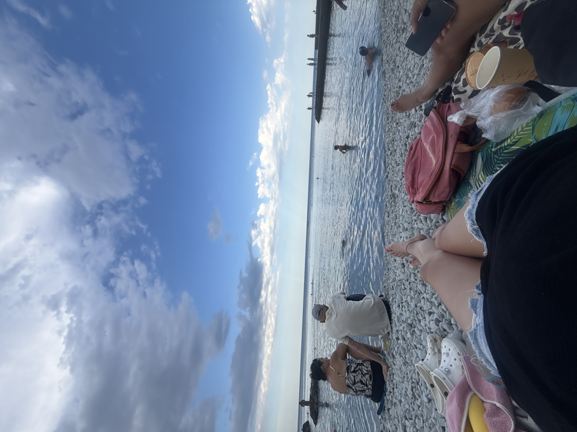 In the city of Partenit, Ukraine, a group of people are enjoying a day at the beach. The scene is captured from a low-angle, first-person perspective, looking out towards the sea. The foreground shows a beach covered in smooth, light-colored pebbles. Scattered on the pebbles are various personal items: a pair of white Crocs, a pink towel with a yellow object peeking out, a pink backpack, a plastic bag containing what appears to be food, and a paper cup. A person's legs, wearing denim shorts and a black top, are visible in the lower portion of the frame. To the right, another person's legs are visible, and they are holding a black smartphone.
Further back, a couple sits on the pebbles facing the water. The woman wears a black and white patterned swimsuit and the man wears a white t-shirt and a grey cap. The sea is calm and reflects the blue sky, which is dotted with white and grey clouds.
In the water, several people are visible. A child stands in the shallow water near the shore. Further out, a person is swimming or floating, and on a long concrete pier or breakwater in the distance, several silhouettes of people are standing. The overall impression is of a relaxed, leisurely day at the seaside.