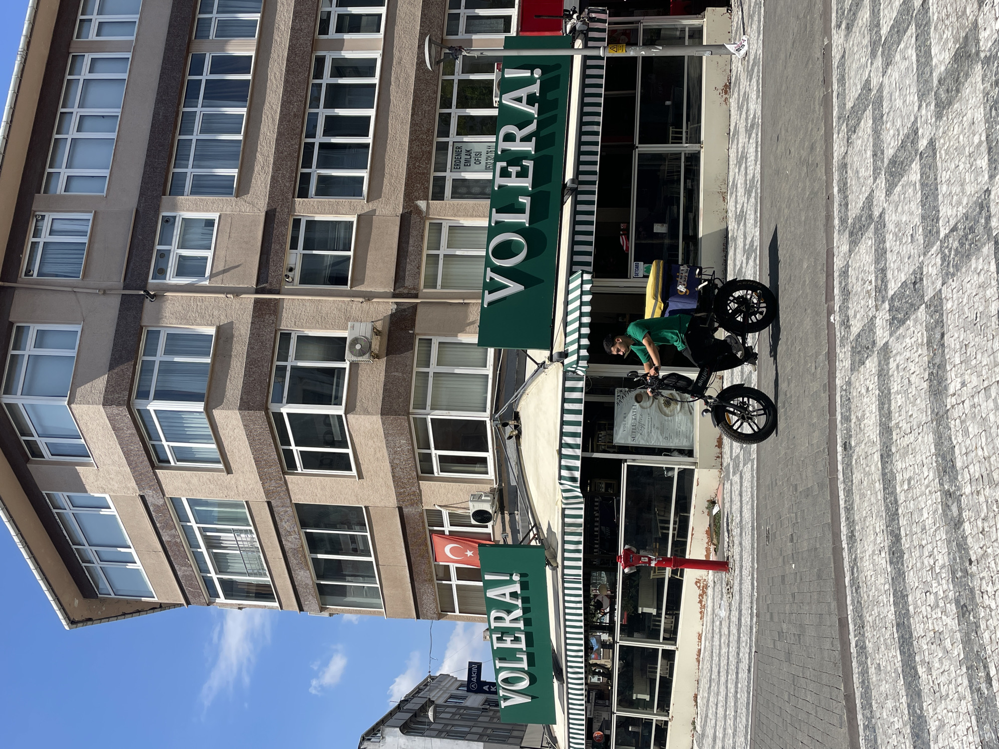 An exterior shot captures a building in Kadıköy, Turkey, on a sunny day. The building has a beige facade with multiple floors of windows. On the ground floor, a restaurant or cafe called "VOLERA!" is visible with a green awning striped with white and green. A man in a green shirt is riding a black electric scooter with a yellow and blue delivery box on the back across a cobblestone street. A red fire hydrant is present on the sidewalk. In the background, a Turkish flag is displayed from the building, and a sign for "ERDENER EMLAK OFİSİ" is visible in a window. The sky is blue with some scattered clouds.
