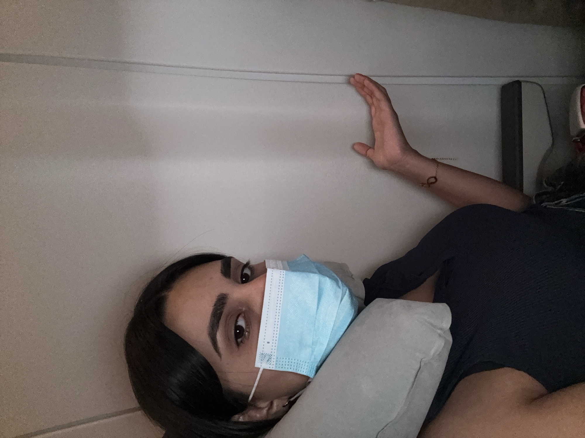 A person wearing a blue surgical mask is lying down, possibly in an enclosed space such as an airplane or a small room. The person is resting their head on a light gray pillow. Their right arm is extended upwards and the hand is touching a light-colored wall. A gold bracelet is visible on the arm. The person is wearing a dark blue or black shirt. The location is not discernible as Geghanist, Armenia. The background consists of plain light colored walls.