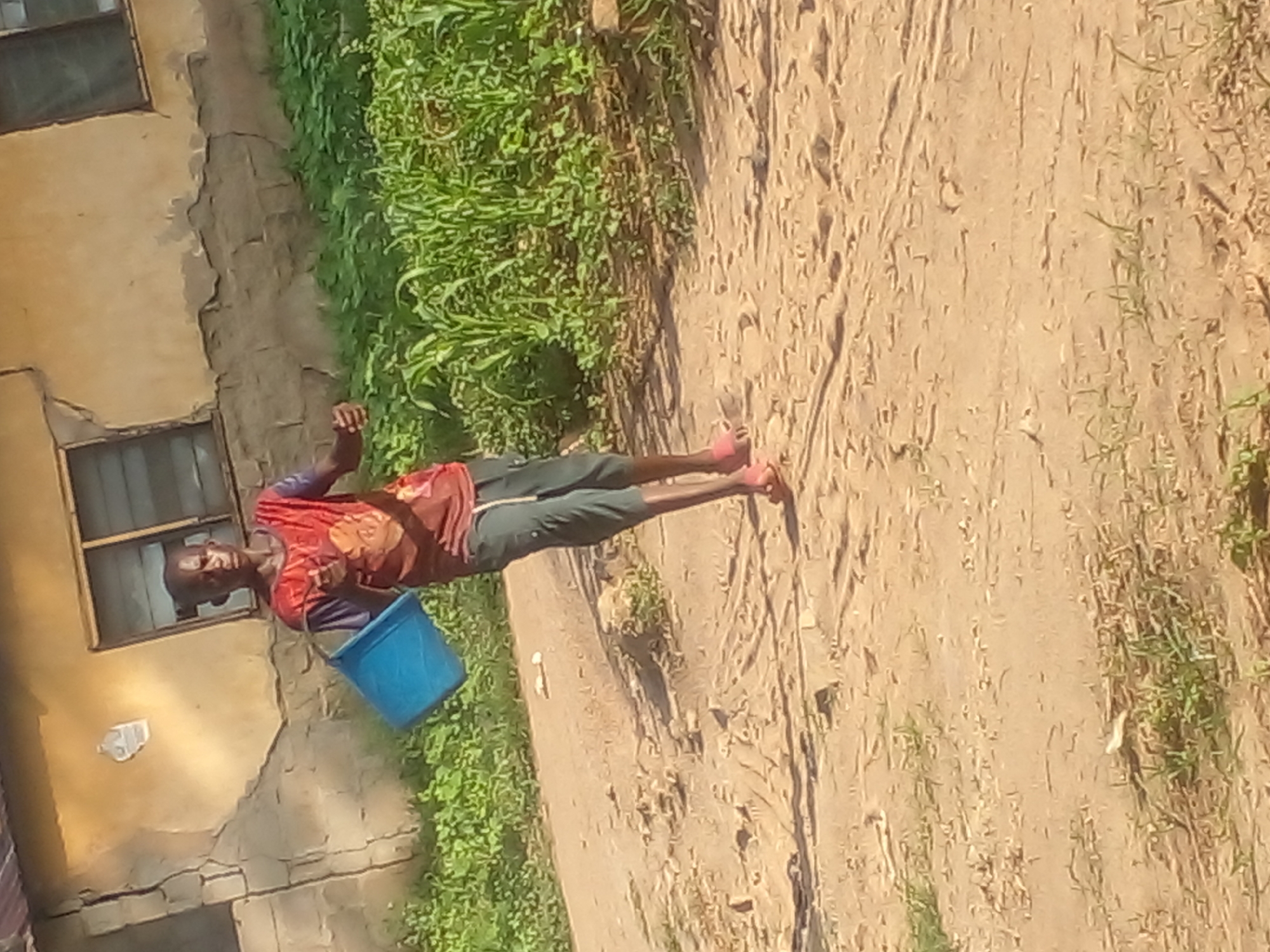 A young Black male is captured in a vertical, slightly tilted photograph. He is standing in an outdoor setting, facing the camera and holding a blue bucket in his left hand. He wears a red t-shirt with a graphic design and dark green pants. His footwear is visible, appearing to be pink or light red.
The background consists of a rough, textured wall on the left, with a window above and another visible lower down. Green vegetation grows along the side of the wall. To the right of the person, the ground is sandy and appears to be a dirt path with some sparse vegetation. The lighting suggests it is daytime, likely sunny, casting shadows. The overall impression is of a rural or semi-rural environment, possibly in a residential area. There is no visible text in the image. The subject's expression is neutral.