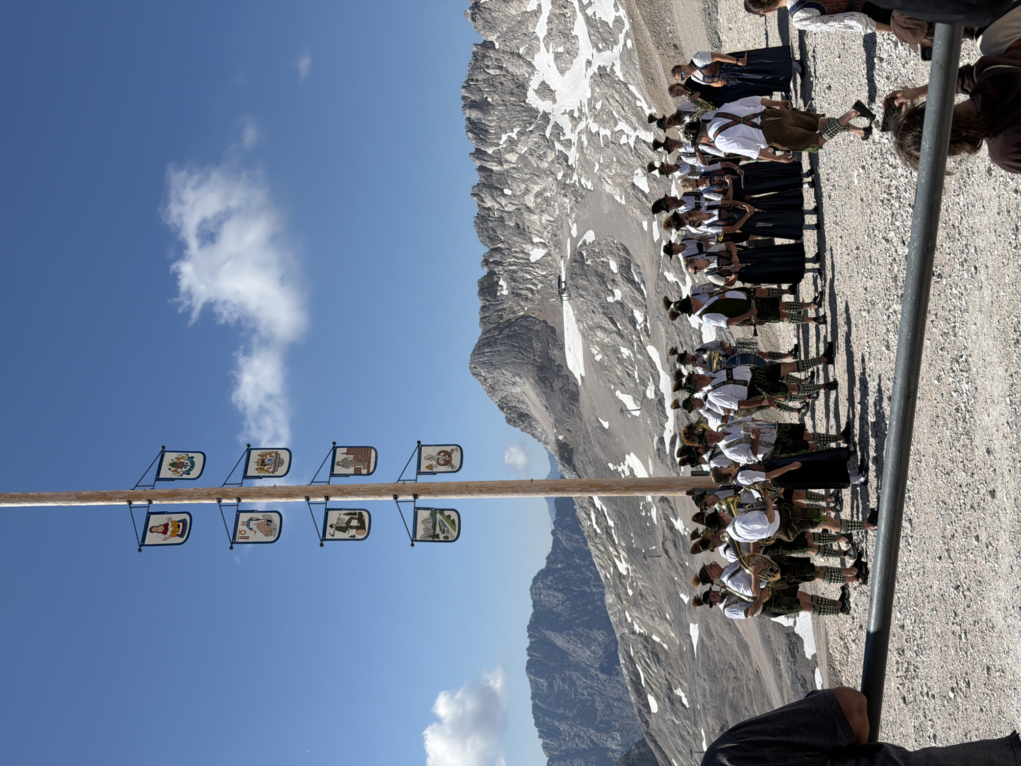 A group of people in traditional Bavarian attire, some playing musical instruments, stand on a gravelly surface in a mountainous region. A tall wooden pole adorned with small banners featuring illustrations stands prominently in the foreground. In the background, rugged, snow-dusted mountains rise under a clear blue sky with scattered clouds. The people appear to be gathered for an event or performance. The scene suggests an outdoor festival or cultural gathering in a high-altitude location. The specific location in Leutasch, Austria, is indicated by the context of the query.