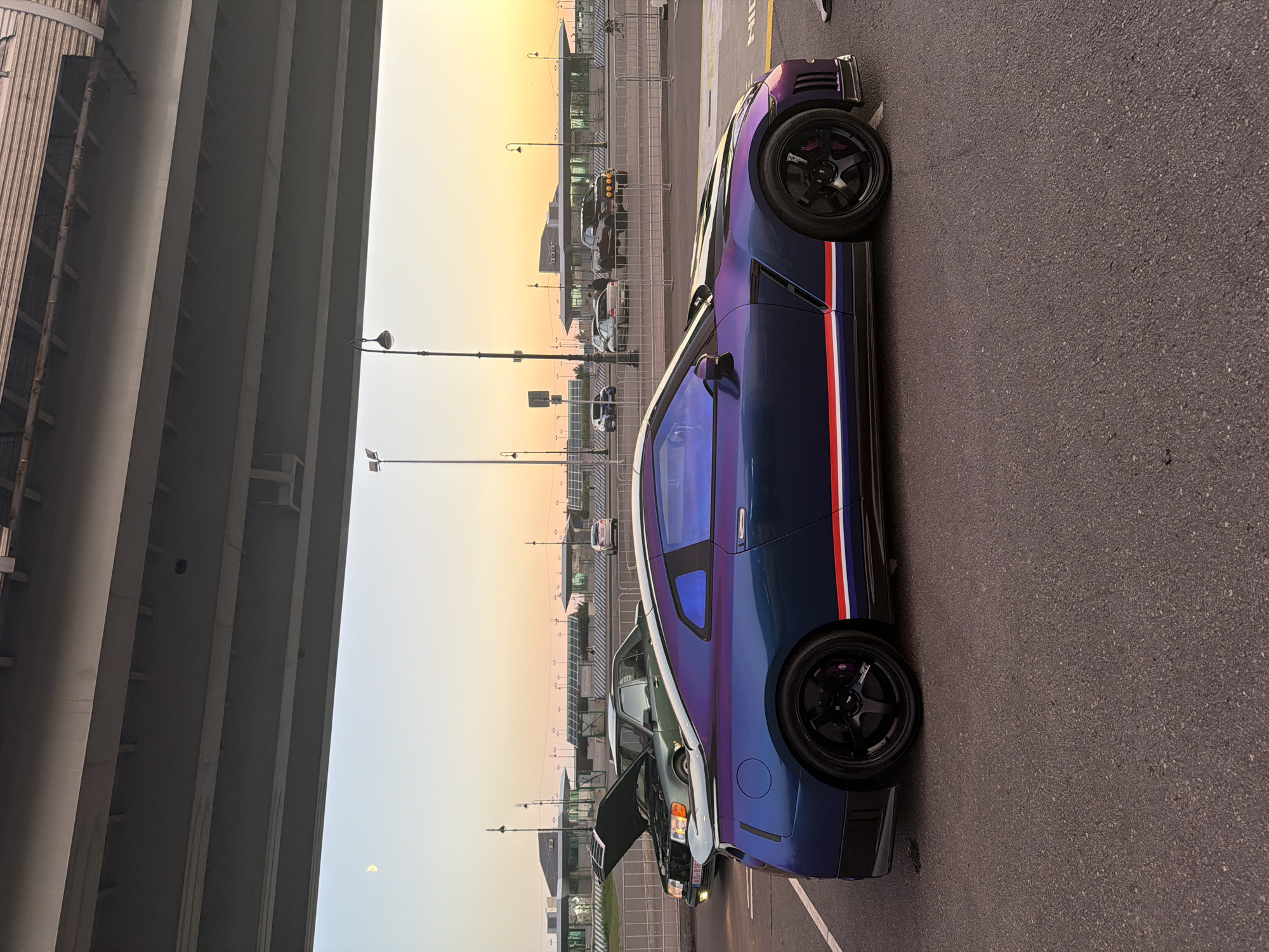 A Nissan GT-R car is parked on asphalt under a highway overpass. The car has a iridescent paint job that shifts from blue to purple and features a red and white stripe along the side. In the background, a vintage green Mercedes-Benz sedan with its hood open is parked. Beyond the cars, there is a modern building with a glass facade and a row of lampposts. The sky is a gradient of light blue to orange, suggesting it is either sunrise or sunset. The moon is visible in the upper left quadrant of the sky.