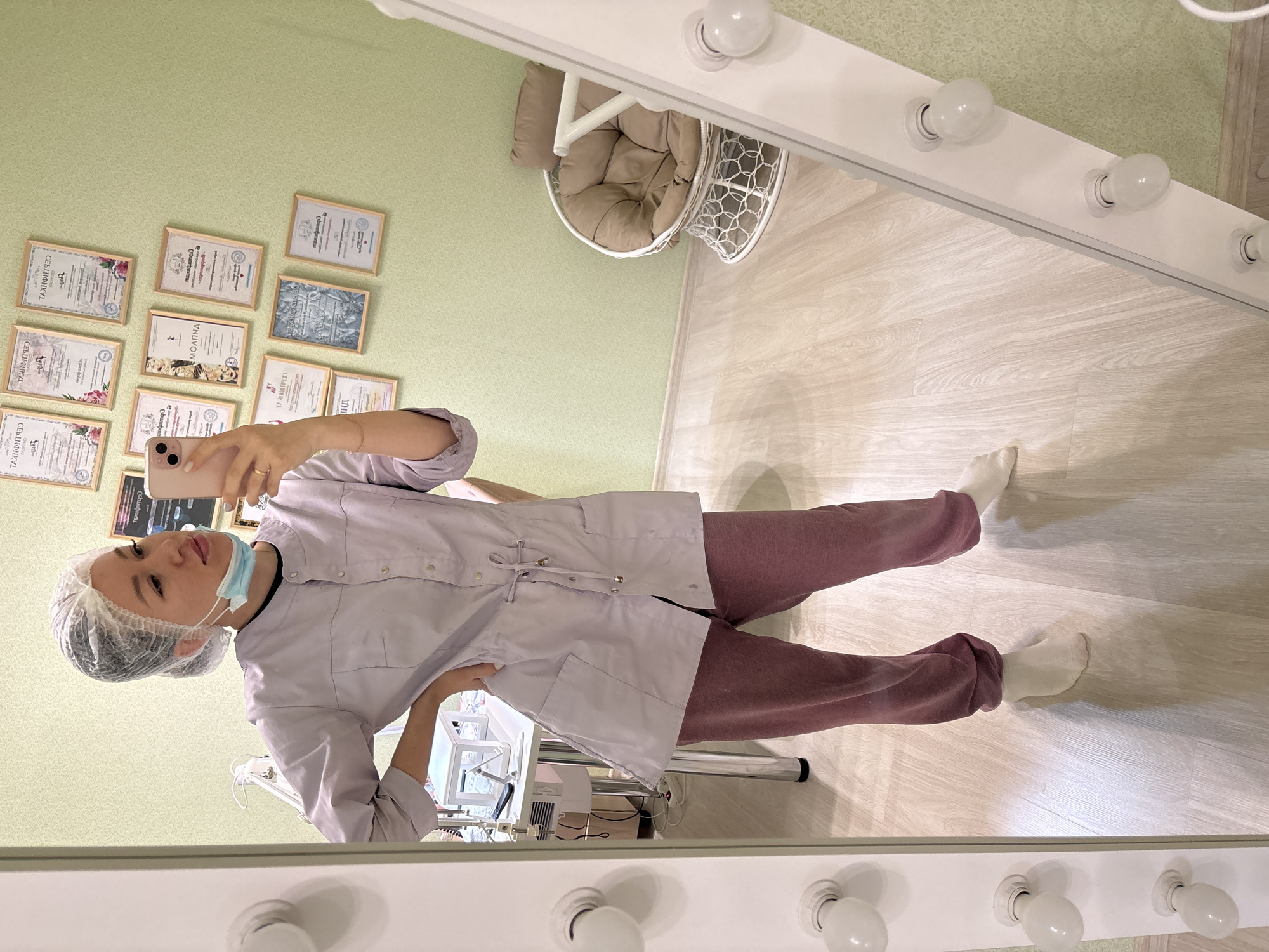 A woman in a light purple lab coat, maroon pants, and white socks stands in a room with light green walls in Kostoryata, Russia. She wears a disposable hair net and a blue surgical mask. She holds a smartphone in her left hand, presumably taking a selfie in a mirror with light bulbs bordering it. Behind her are framed certificates on the wall, a white wire chair with a tan cushion, and a medical device with cords. The floor is light-colored wood.