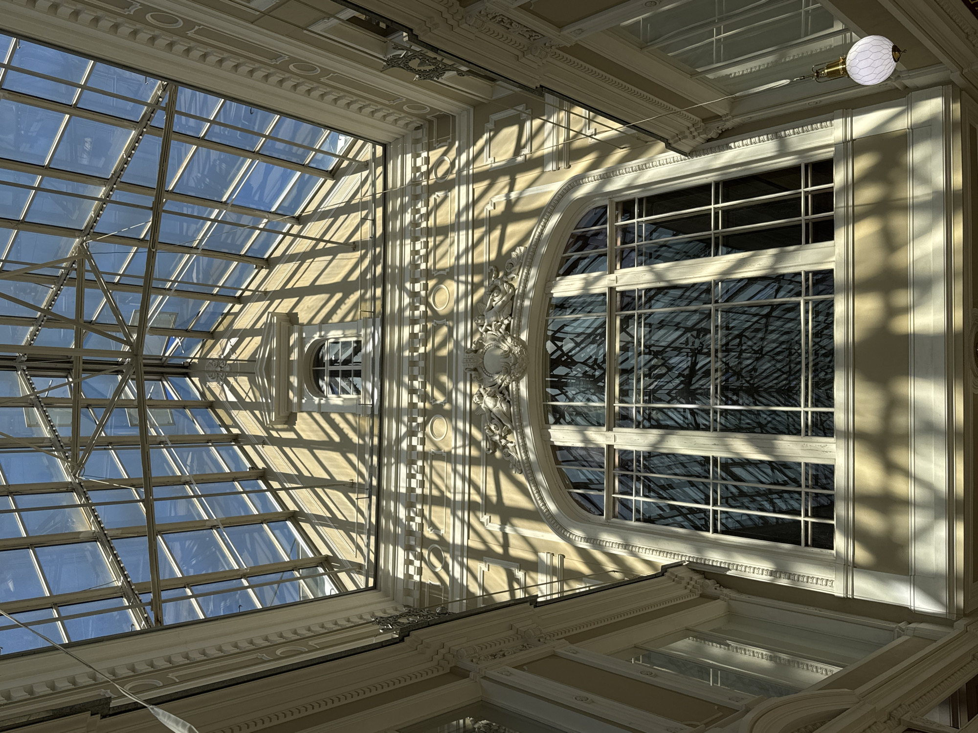 The image shows an interior view of a building in Saint Petersburg, Russia, looking upwards towards a glass ceiling. The architecture is ornate with decorative moldings and classical detailing. Sunlight streams through the gridded glass of the ceiling and windows, creating patterns of light and shadow on the walls. A hanging light fixture is visible in the upper right corner. The overall tone is bright and airy.