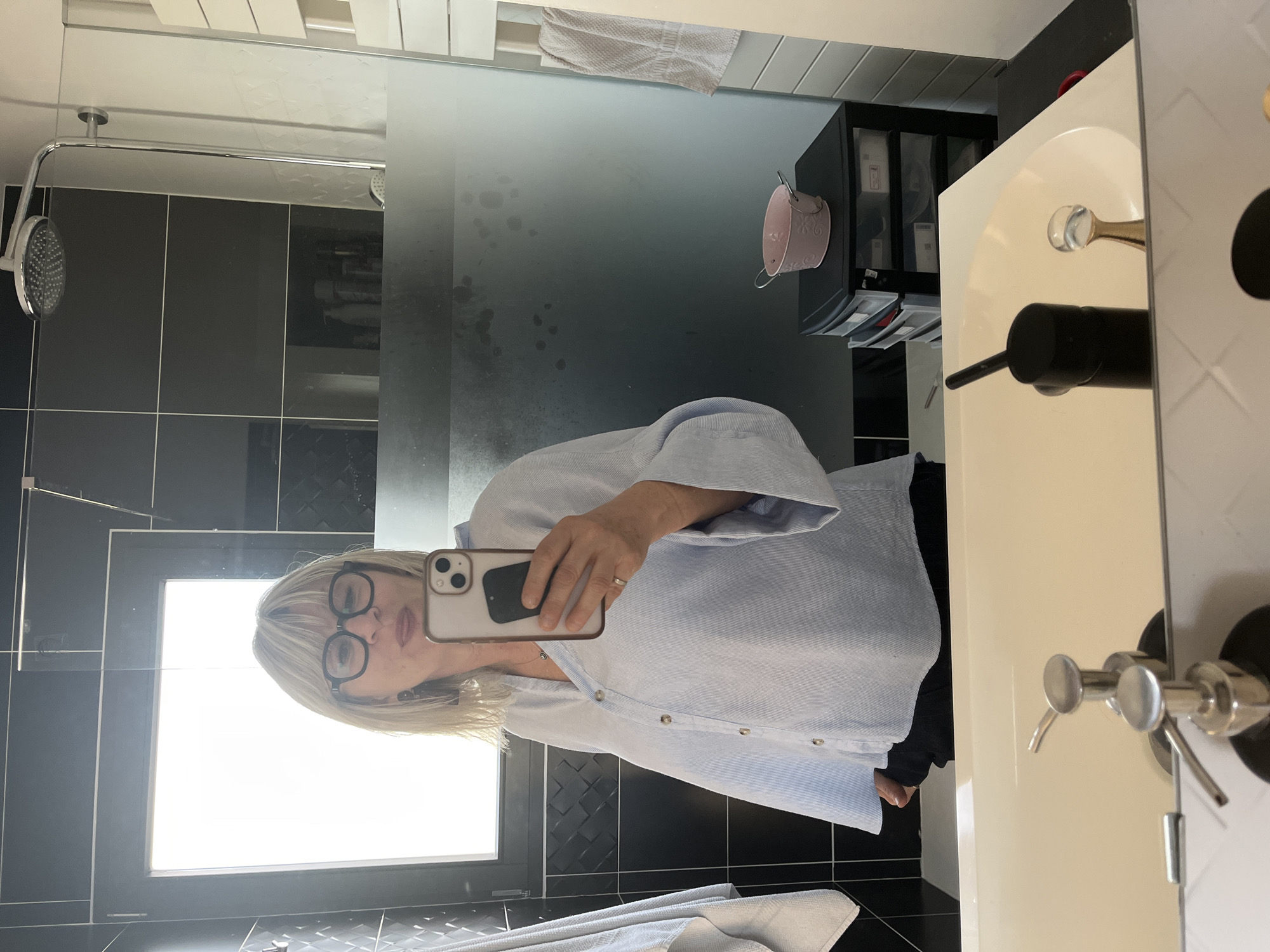 A selfie taken in a bathroom. The bathroom features black and white checkered tiles, a shower with a glass door, a towel rack with a white towel, and a white sink. A woman wearing glasses and a light blue shirt is taking the photo in the mirror. The location is Chambœuf, France.