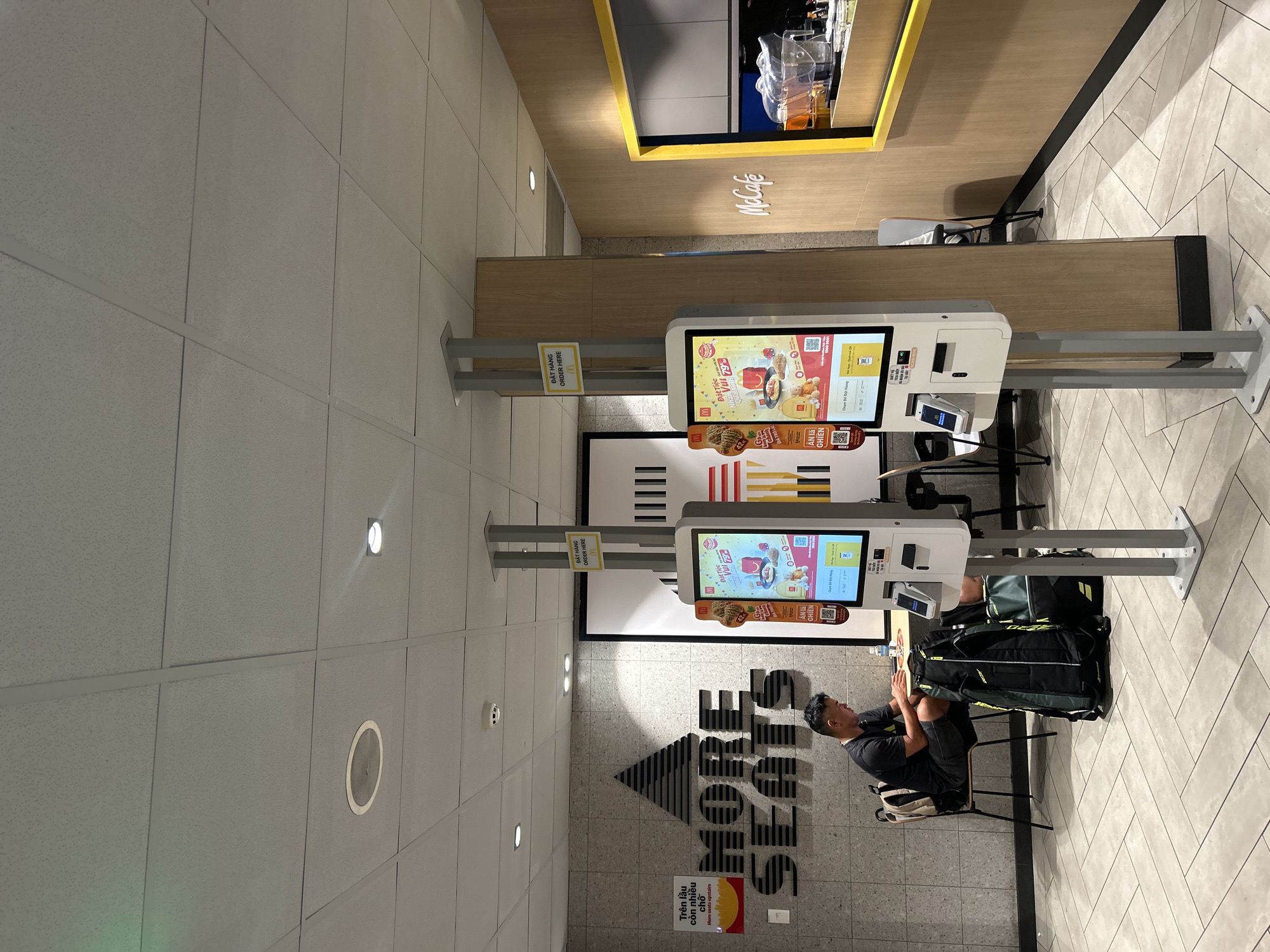 The image depicts the interior of a McDonald's restaurant in Thủ Đức, Vietnam. Two self-order kiosks stand prominently in the foreground. A man is seated at a table, facing a tablet, with luggage bags next to him. Behind him, a large "MORE SEATS" sign is visible on a tiled wall. The restaurant has a modern, clean aesthetic with a suspended ceiling and wood-paneled walls. The McCafe branding is visible on the right side of the image.