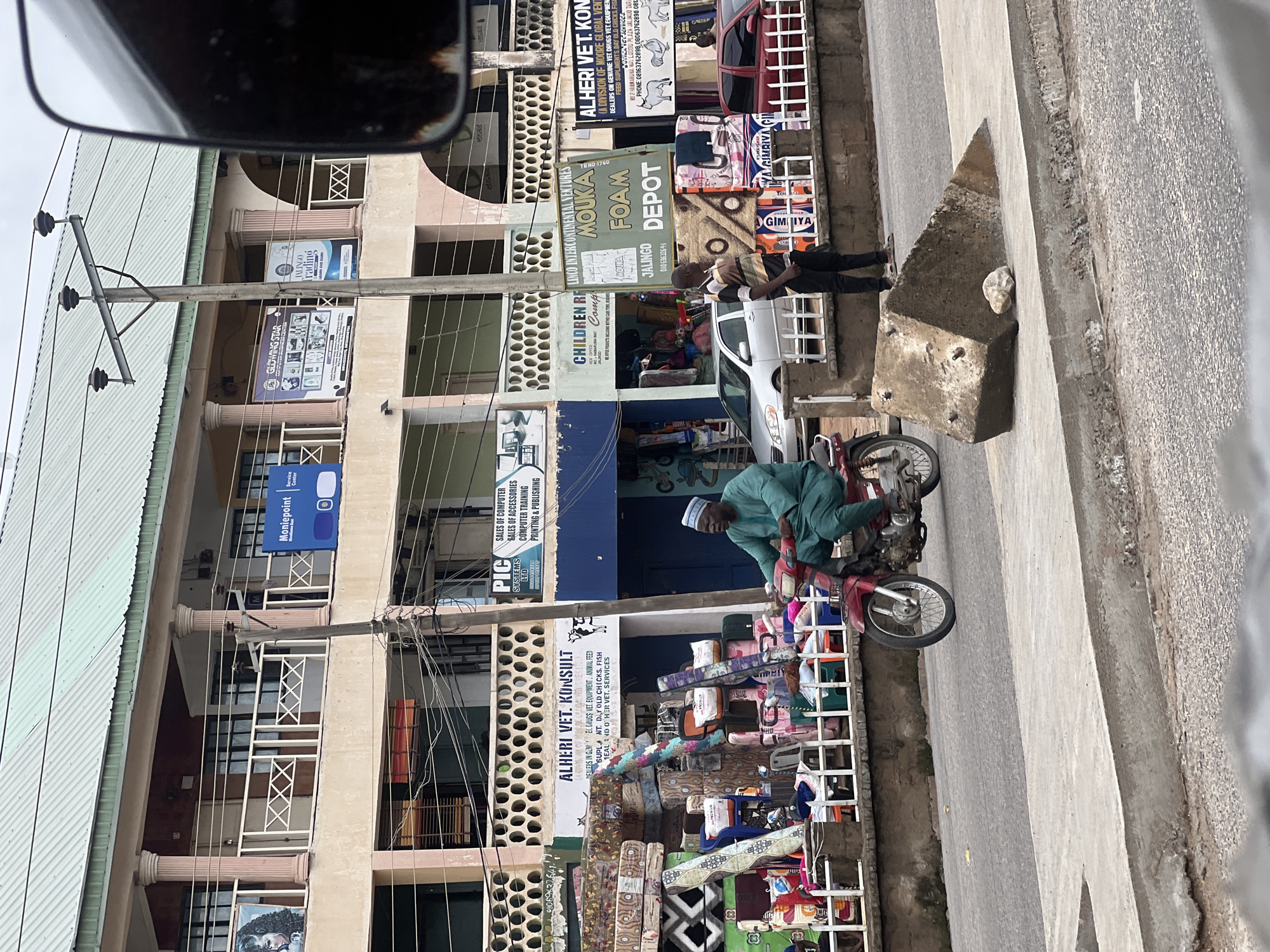 The image captures a street scene in Jalingo, Nigeria. In the foreground, a man in a green traditional outfit rides a red motorcycle. The motorcycle is on a paved road. To the right of the motorcycle, a man in a striped shirt and dark pants stands on the sidewalk. Behind the man on the sidewalk, a large concrete barrier is present on the sidewalk. A white railing separates the sidewalk from a storefront display. The storefront features stacks of mattresses and rolls of fabric. Above the storefront, a multi-story building has shops on the ground floor. Signs for "ALHERI VET. KONSULT," "PIC SYSTEMS LTD," and "CHILDREN COMP..." are visible. Electrical poles and wires are visible overhead. The sky is overcast.