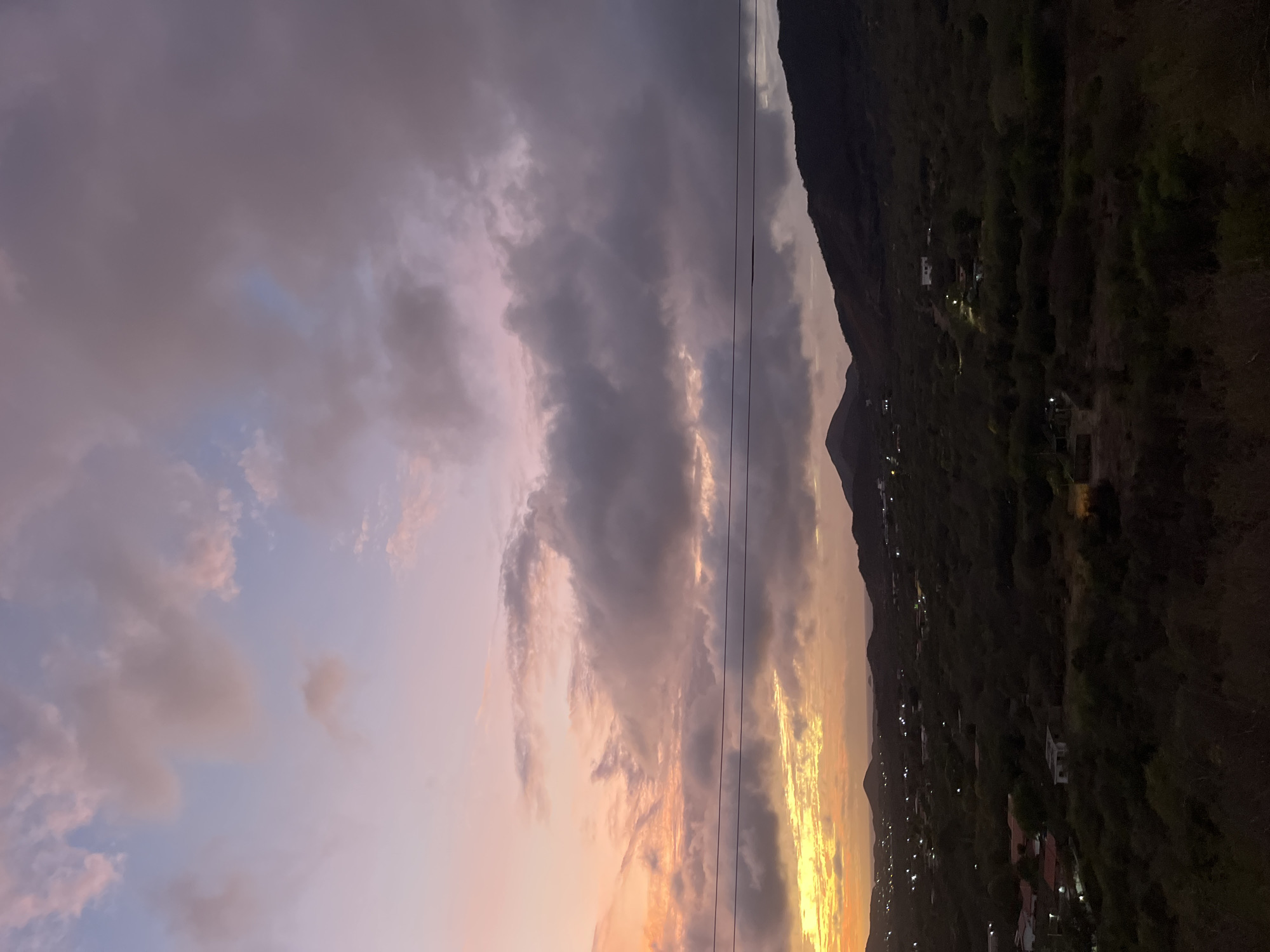 The image depicts a twilight landscape, likely from an elevated viewpoint overlooking a valley. The sky is filled with a dramatic display of clouds, transitioning from soft pinks and oranges near the horizon to muted purples and grays higher up. A thin band of bright yellow light suggests the last remnants of sunset.
Below the sky, rolling hills covered in dense vegetation dominate the middle ground. Scattered lights from houses and structures are visible across the valley floor, indicating a populated area. The foreground is a dark, textured expanse of vegetation. Two dark, horizontal lines, likely power lines, stretch across the upper portion of the scene.
The specific location within Santa Ana, Venezuela, cannot be determined from the image alone. However, the scene suggests a semi-rural or suburban setting within a hilly region, characteristic of many areas surrounding cities. There are no people or specific actions depicted. The environment is characterized by natural landscape elements and human habitation.